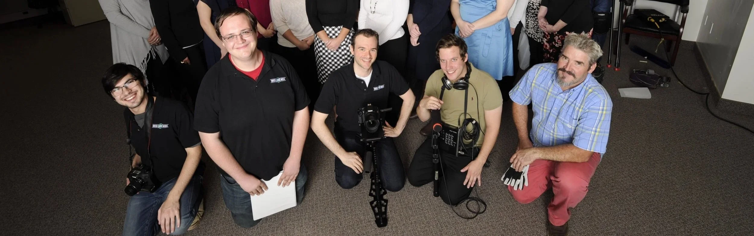 Group photo of a film crew posing indoors with cameras, audio equipment, and a clapperboard.