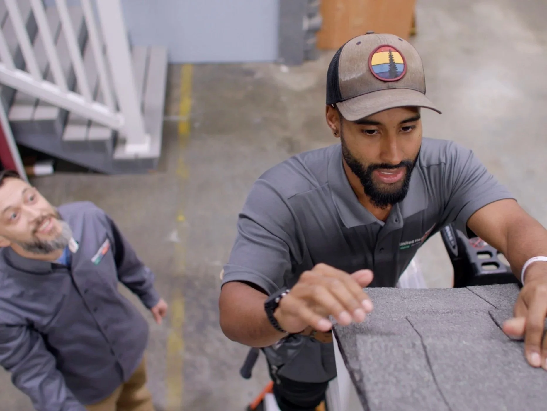 Two workers in gray uniforms loading or moving a large upholstered item in a warehouse