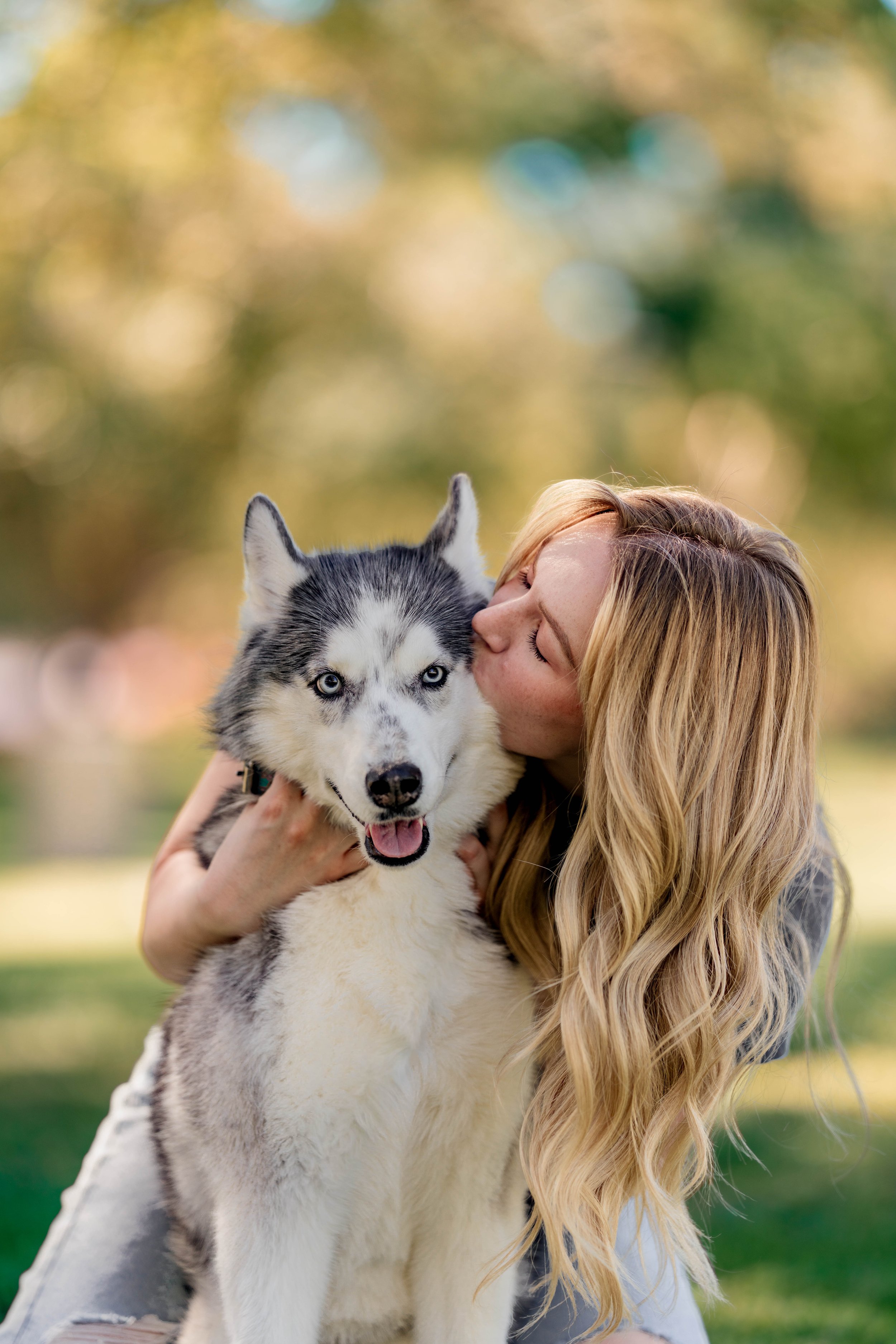 A woman with long blonde hair hugging and kissing a Siberian Husky dog outdoors in a park with blurred trees and green grass in the background.
