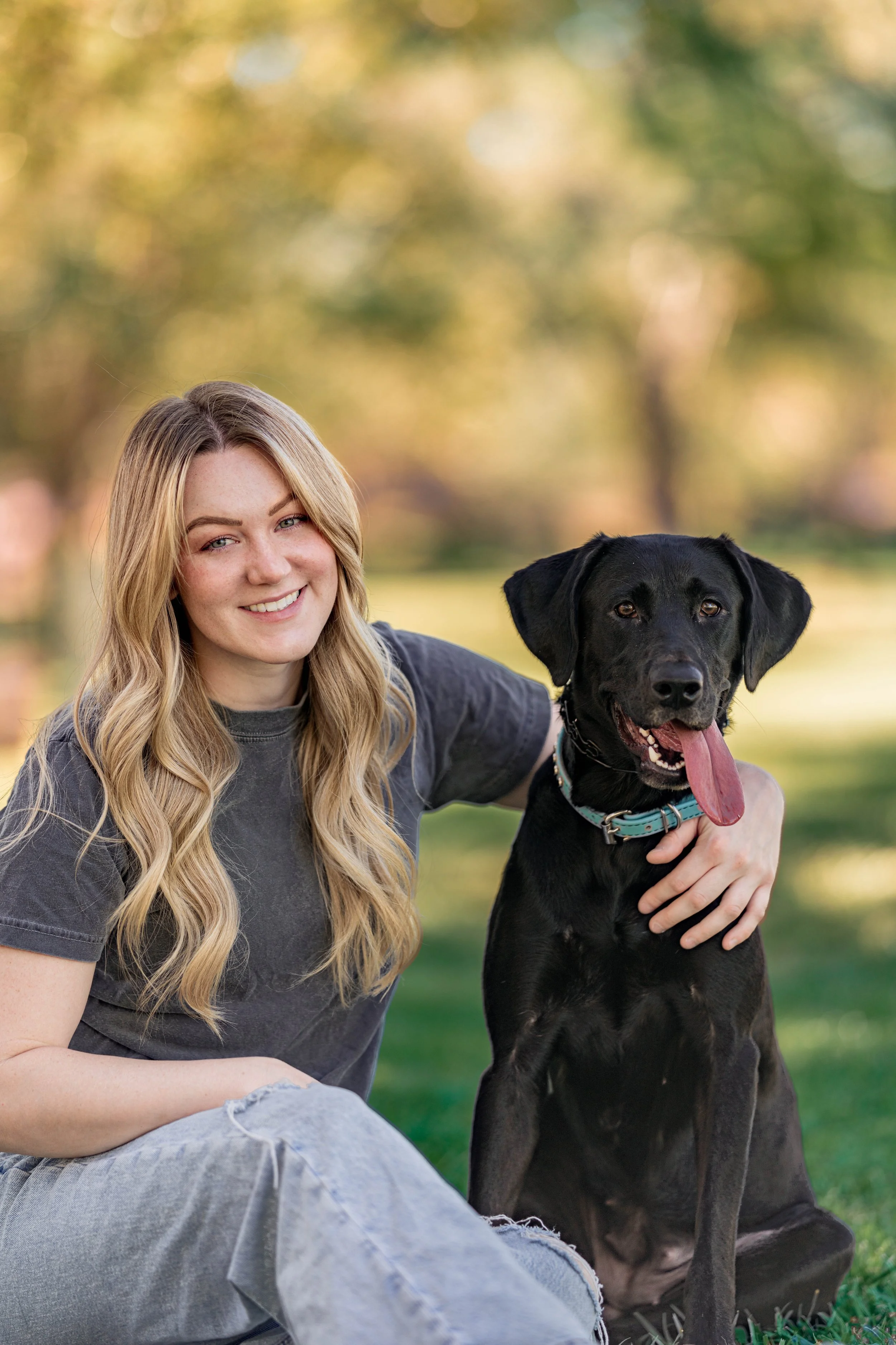 A young woman with long blonde hair and a big smile sitting outdoors with a black Labrador Retriever dog that has its tongue out, in a park with blurred autumn trees in the background.