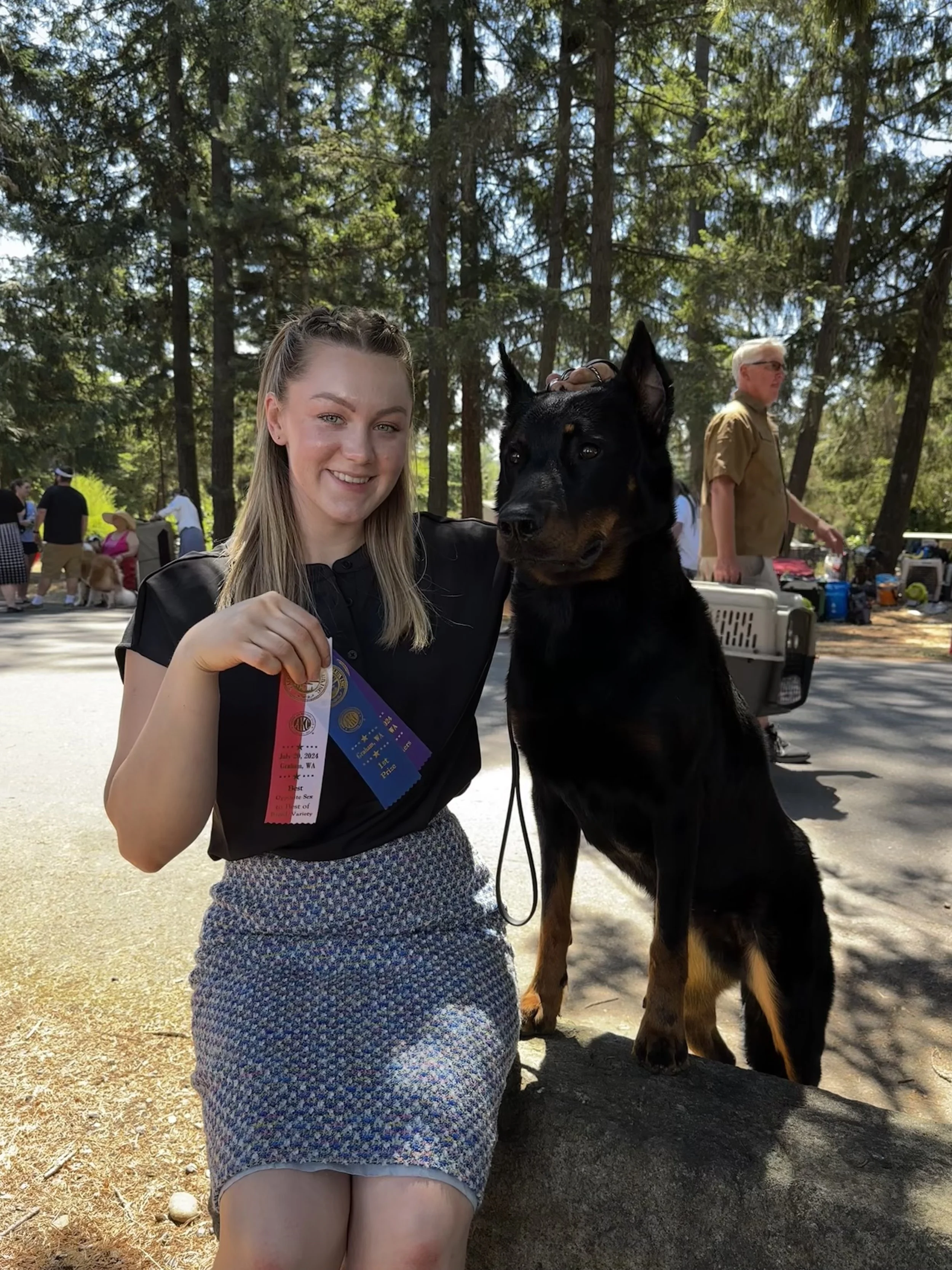 A young woman with blonde hair smiling and holding a ribbon, standing next to a large black and brown dog at an outdoor event in a wooded area. Other people and dogs are visible in the background.