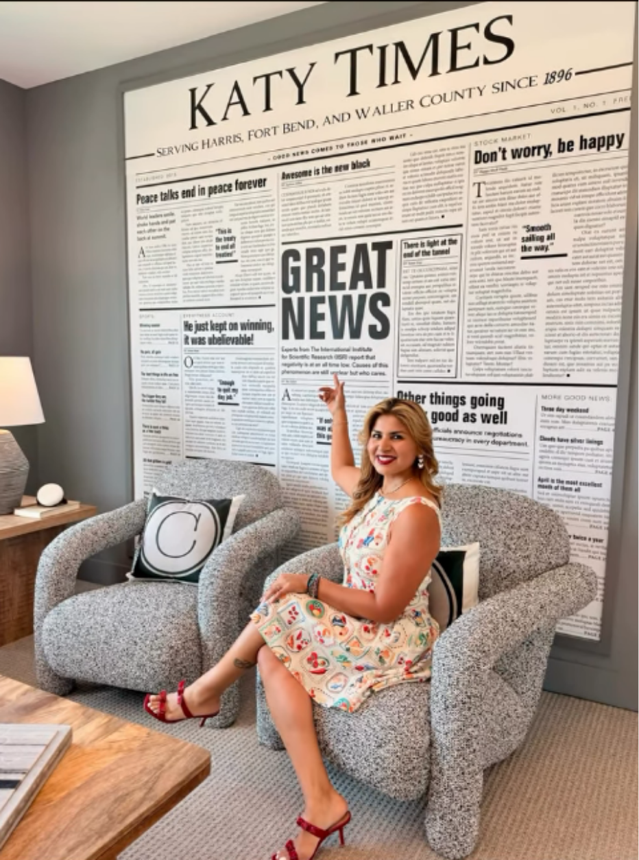 Woman in a floral dress and red heels sitting on a gray armchair, smiling and pointing at a large wall-sized newspaper display titled 'Katy Times' behind her, with a contemporary living room setting.