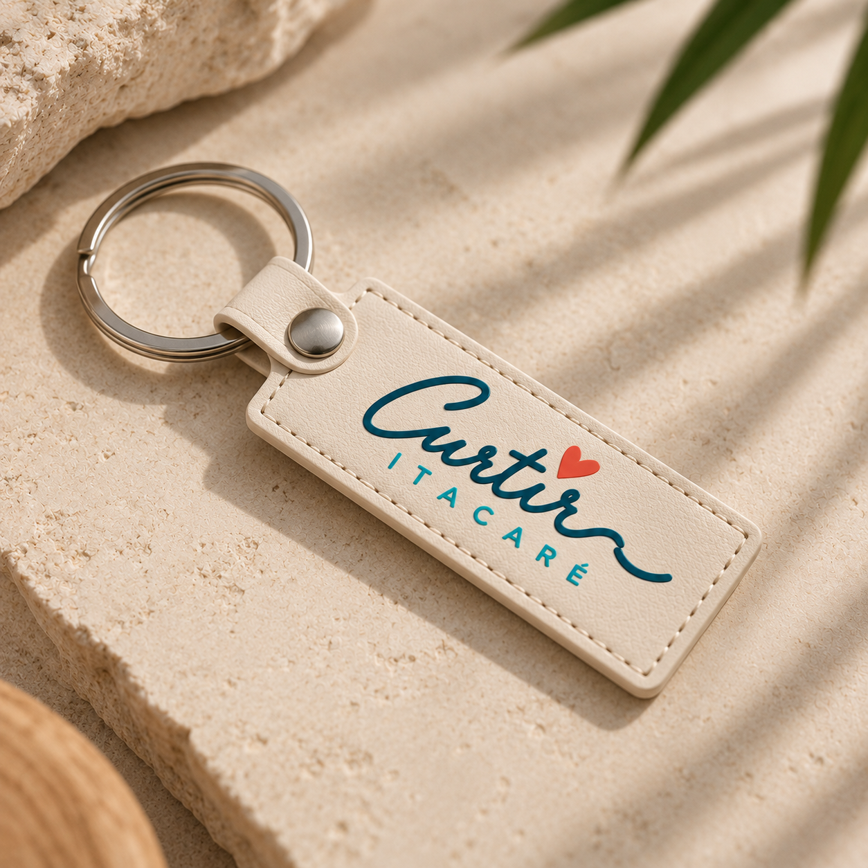 Chaveiro de couro branco com o nome "Cuter Itacaré" escrito em letras azuis, com um coração vermelho no topo da palavra "Cuter", sobre uma superfície de pedra clara, com sombras e folhas verdes parcialmente visíveis.
