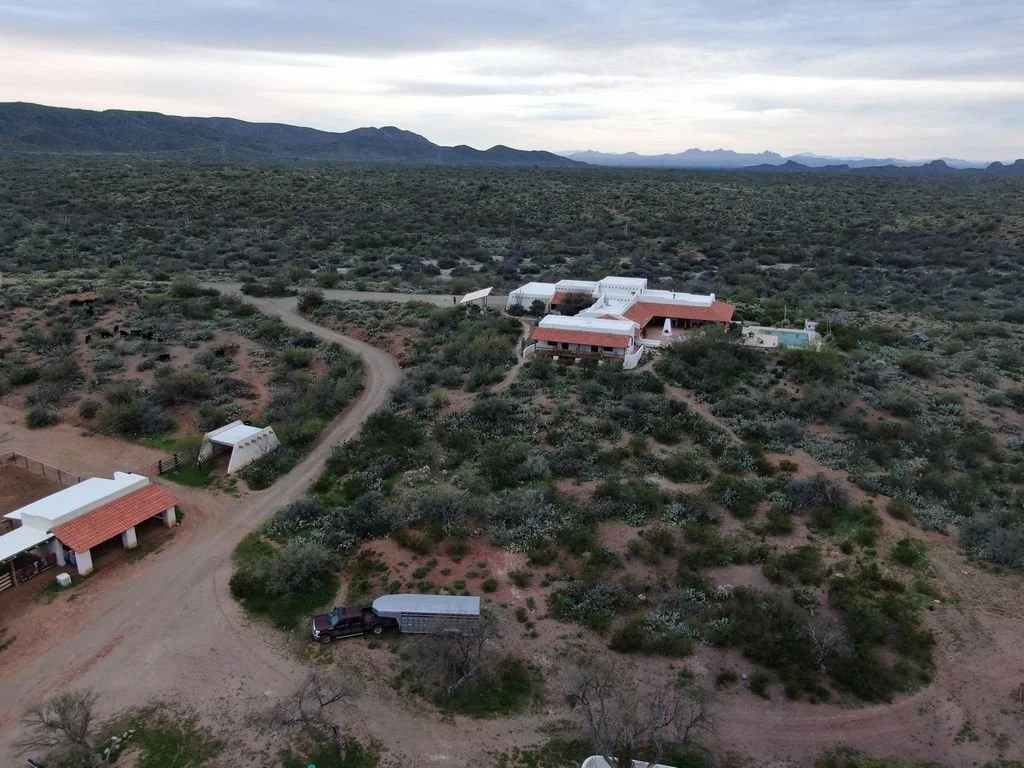 Aerial view of a ranch or farm in a semi-arid landscape with several buildings, dirt roads, and sparse vegetation, with mountains in the background.