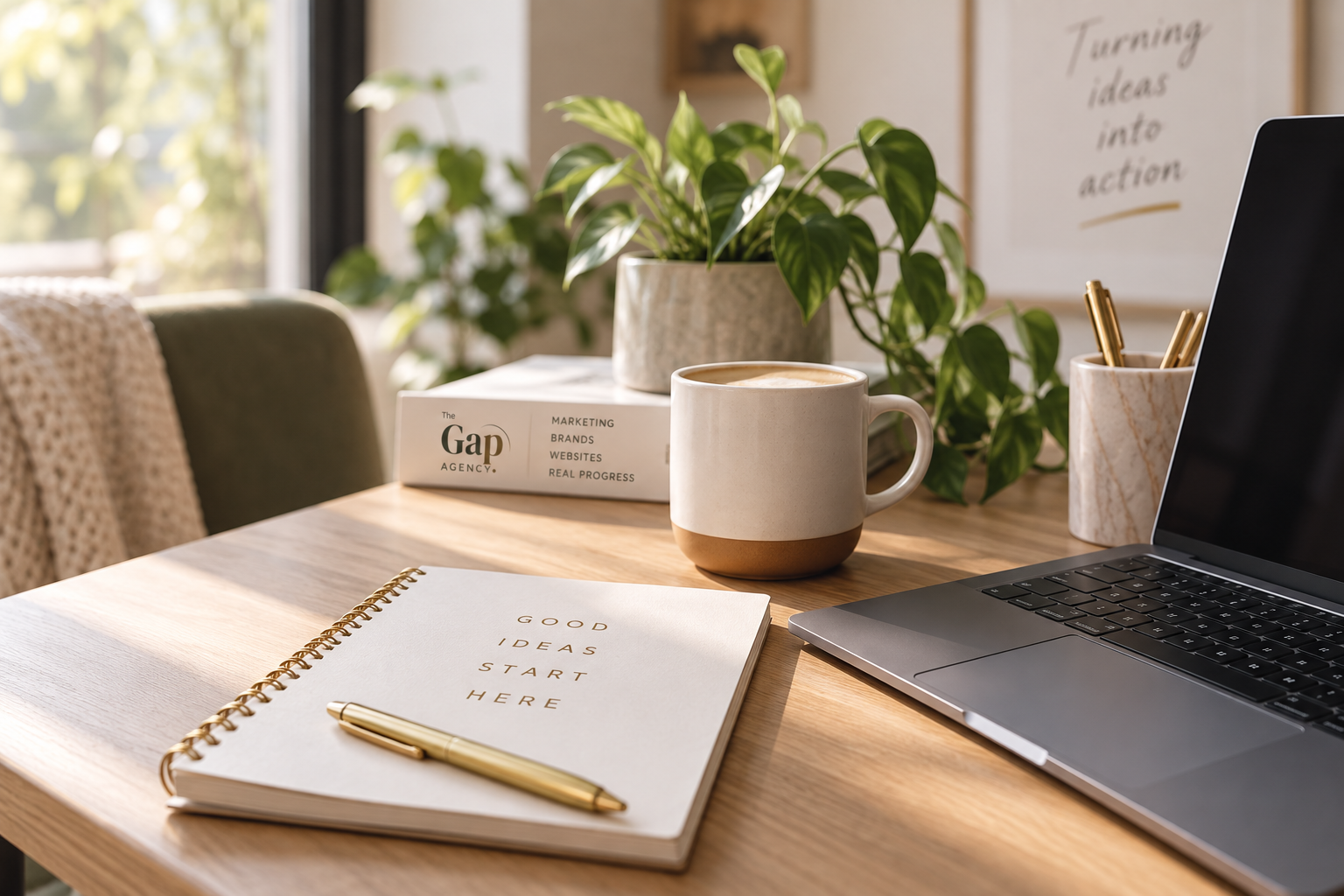 A tidy workspace with a laptop, a coffee mug, a notebook that reads 'Good Ideas Start Here', a gold pen, a box labeled 'The Gap Agency' with marketing topics, a potted plant, and a marble container with gold pens, near a window with sunlight.