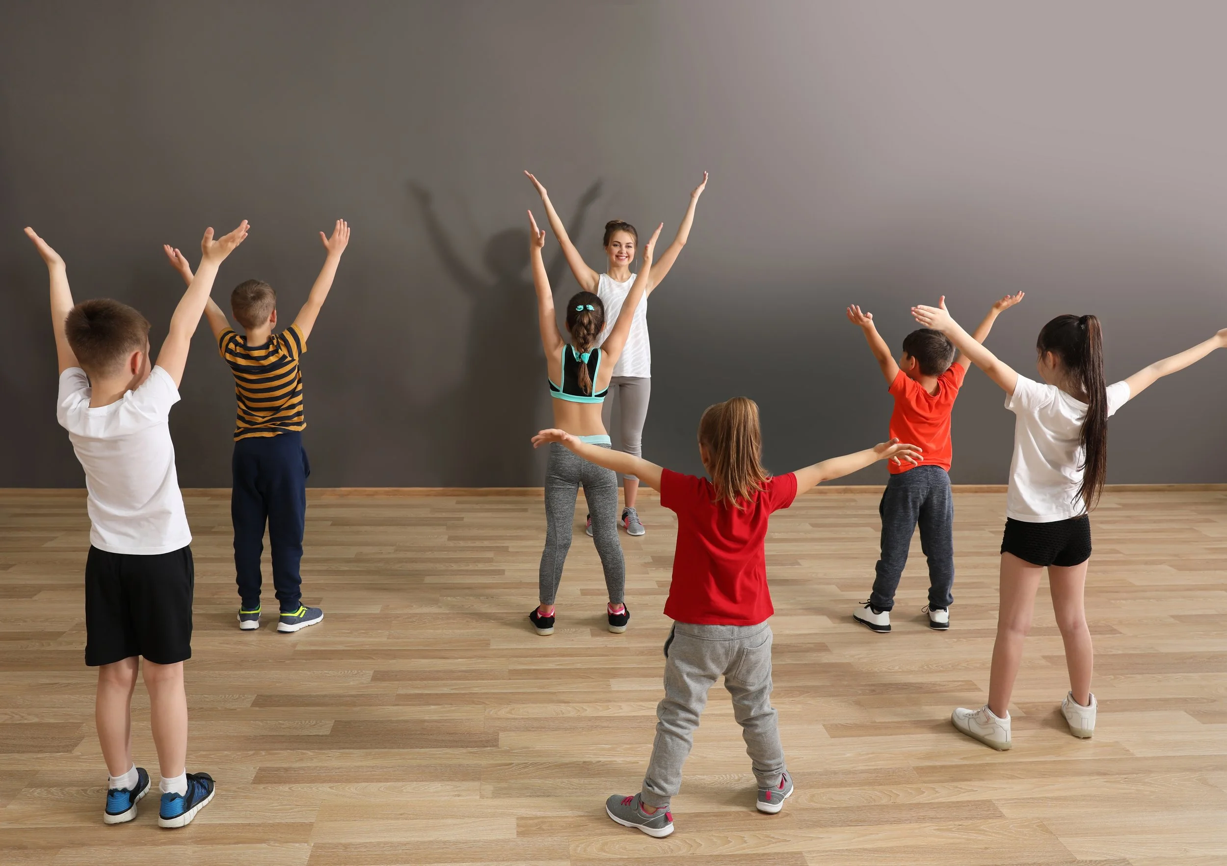 Children participating in a dance or exercise class with an instructor in a studio with gray walls and wooden floors.