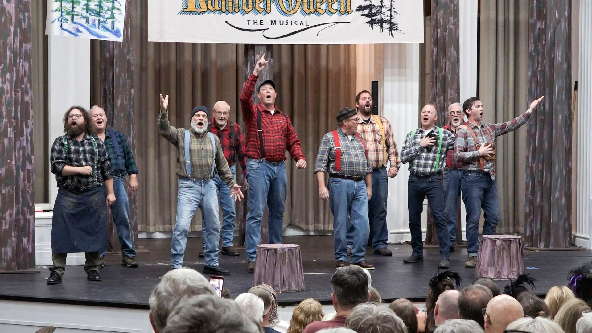 A theatrical performance of 'Muskegon Lumber Queen - The Musical' showing a group of men singing or acting on stage, dressed in plaid shirts and suspenders, in front of a curtain and a banner with trees and text.