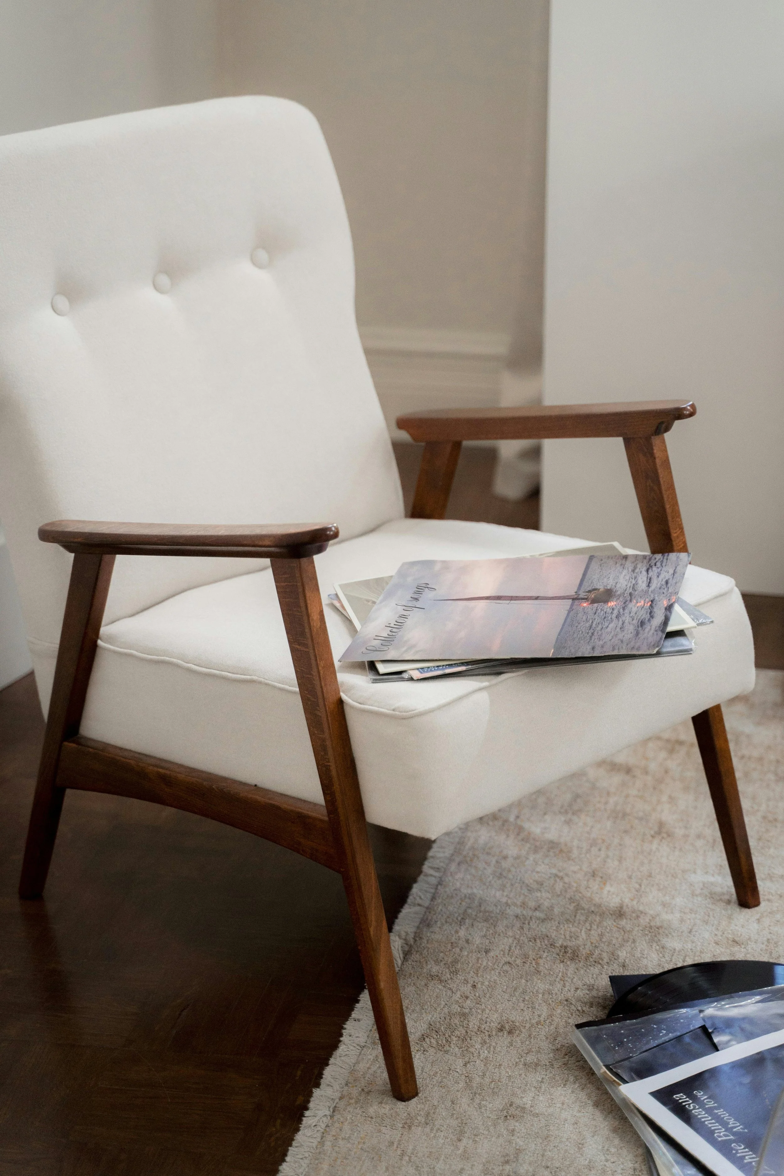 A white armchair with wooden armrests and legs, placed on a beige rug, with magazines or books resting on its seat. Part of a darker wooden floor and a beige carpet are visible.