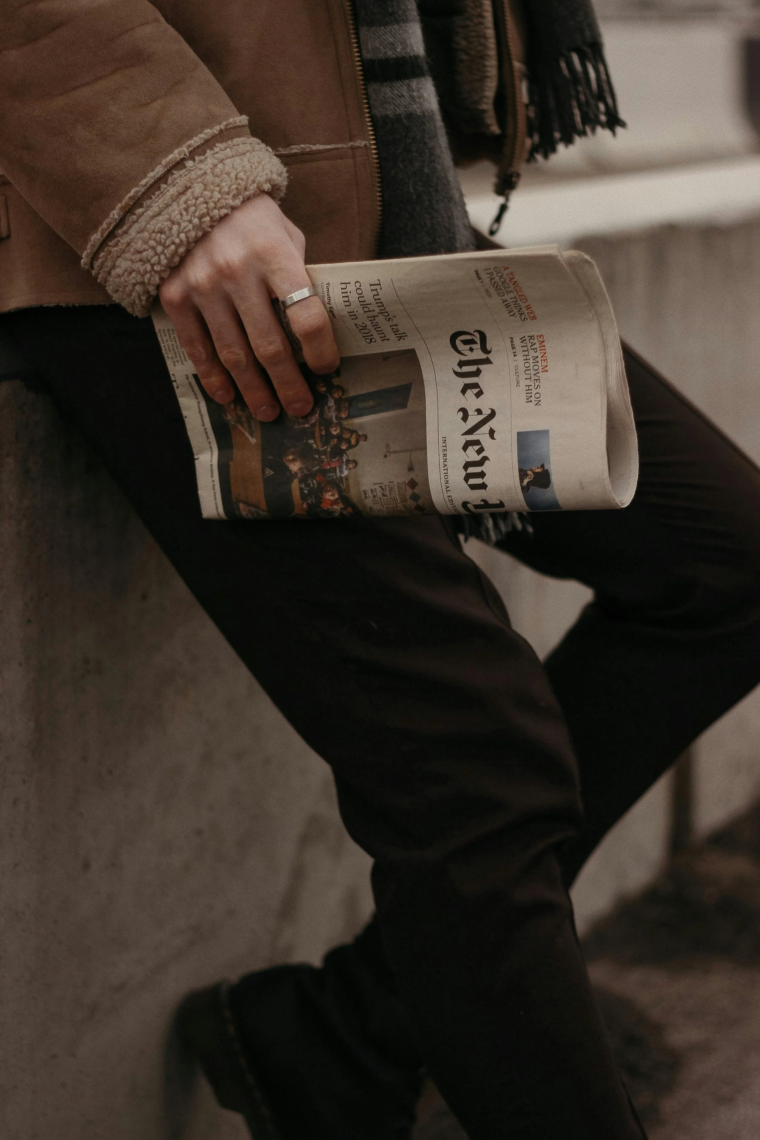 A person holding a folded newspaper in their right hand, wearing a silver ring on their ring finger, a brown jacket with a fleece lining on the cuff, and dark pants, sitting on a light-colored carpeted floor.