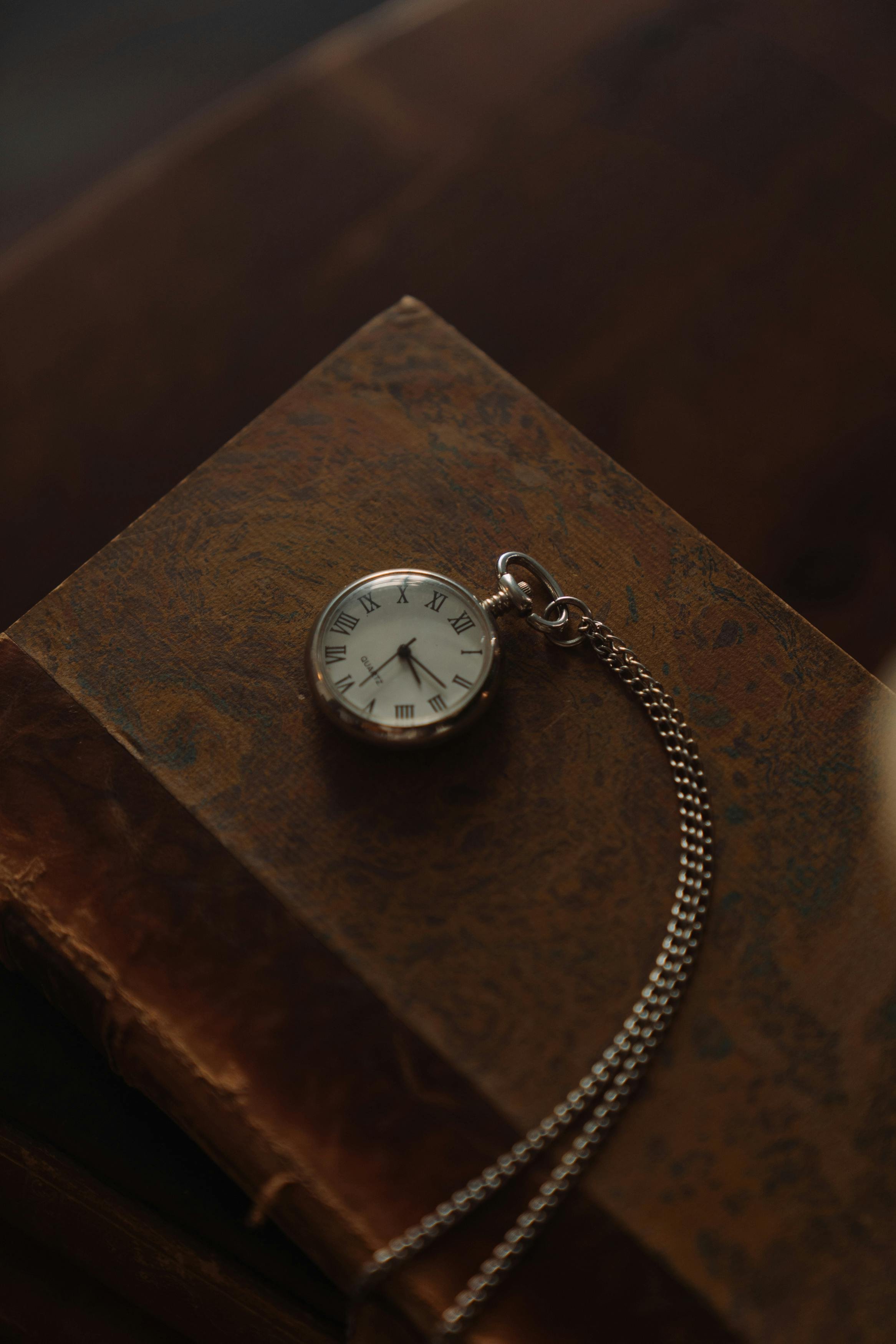 A pocket watch with Roman numerals on a chain, resting on an aged book with a textured, brown cover.