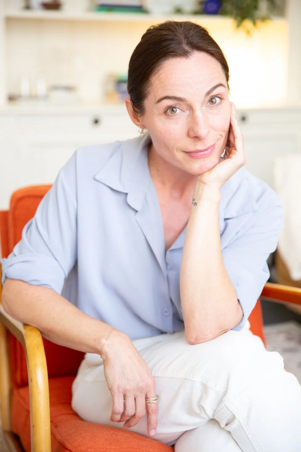 A woman with dark brown hair pulled back, wearing a light blue shirt and white pants, sitting on an orange chair indoors with a thoughtful expression, resting her face on her hand.