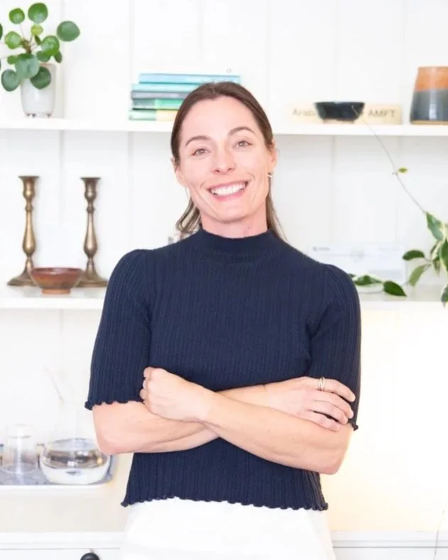 A smiling woman with brown hair wearing a navy blue sweater, standing with arms crossed in a bright room with white shelves, plants, and decorative items.