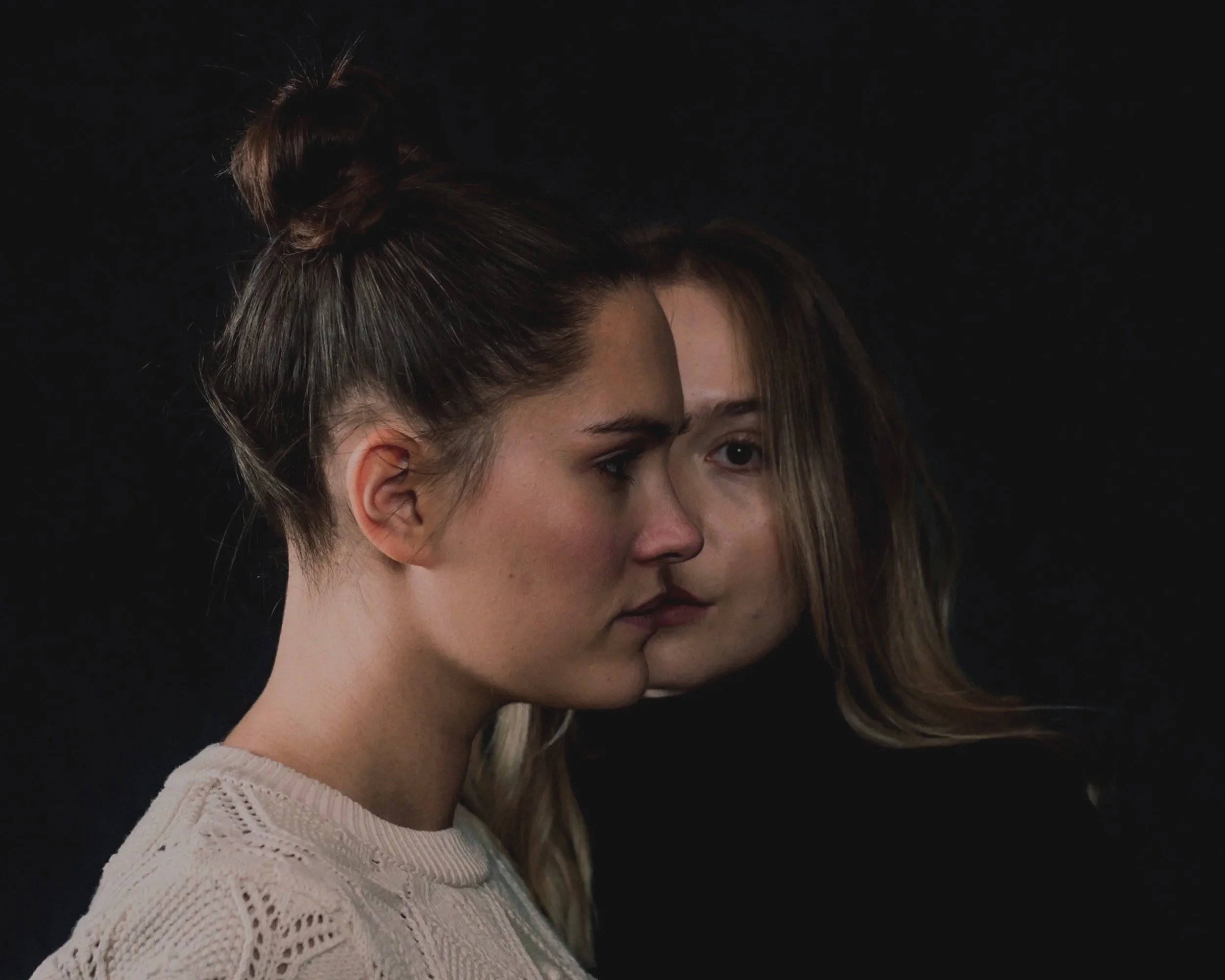 Close-up of two young women kissing each other, with one woman facing left and the other facing right, both with long hair and a black background.