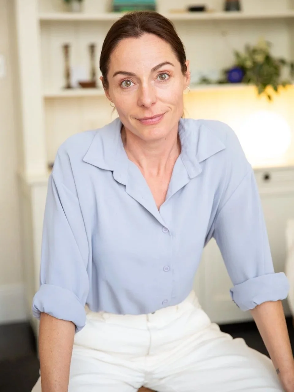 A woman with dark brown hair and fair skin sits casually, wearing a light blue button-up shirt with rolled-up sleeves. She is in a cozy, well-lit room with a bookshelf and decorative items in the background.