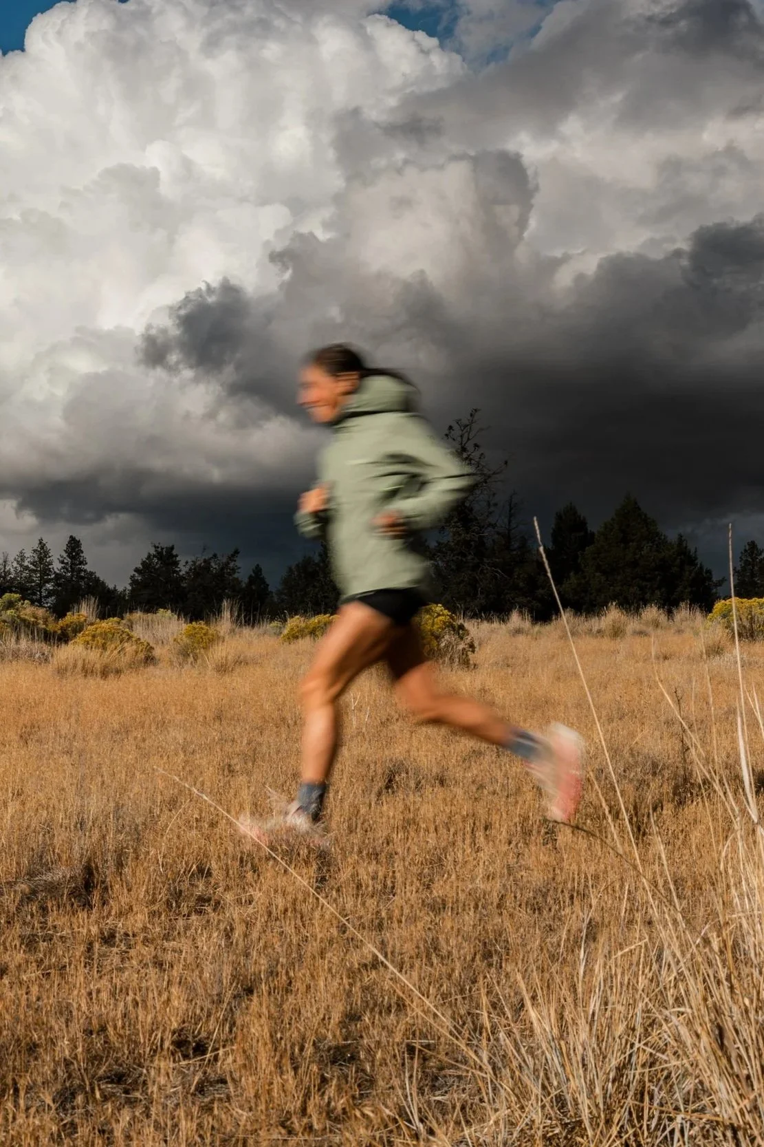 A person running through a grassy field with dark storm clouds and trees in the background.