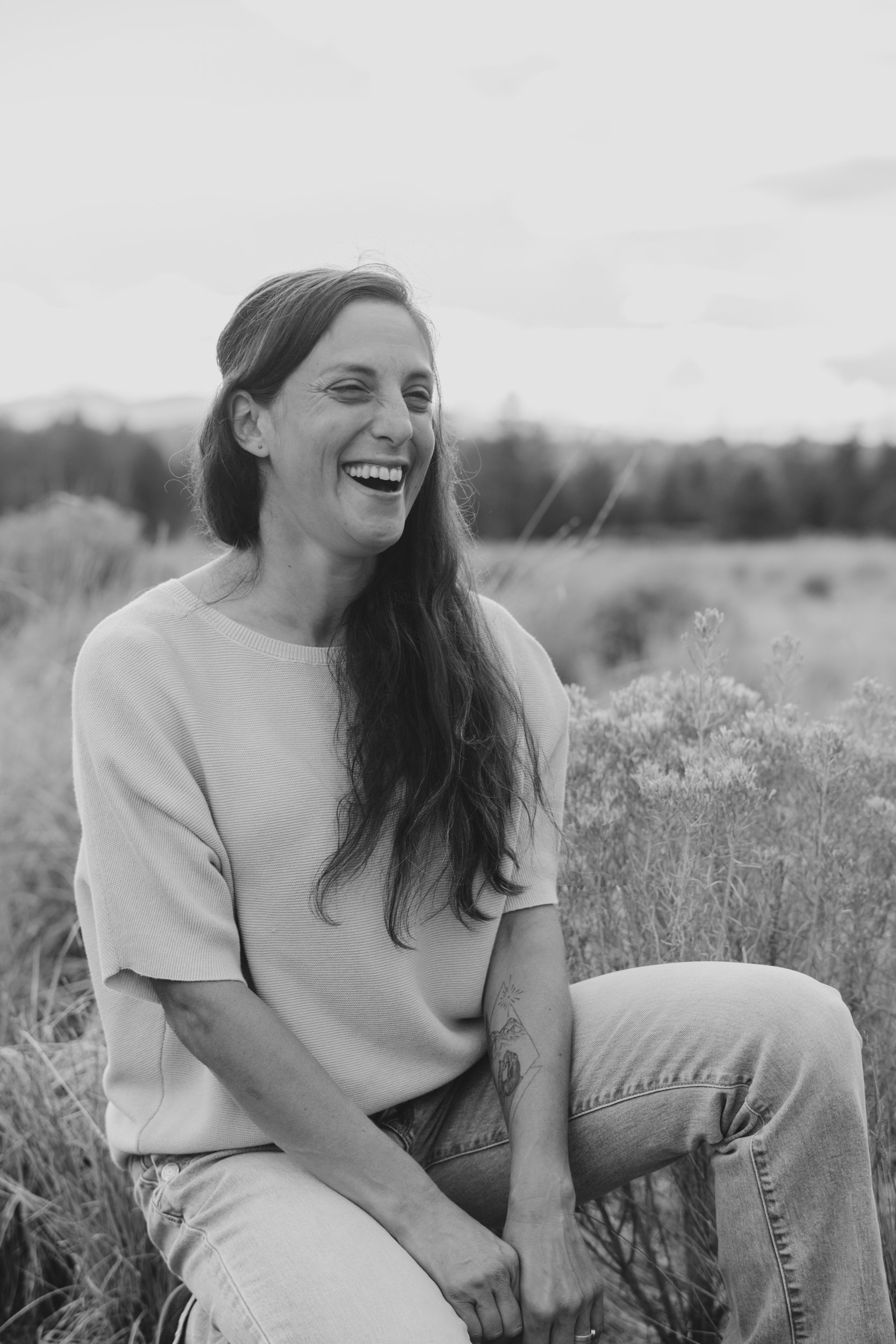 A woman with long dark hair, smiling and looking to the side, sitting outside in a field with tall grass and plants, black and white photo.