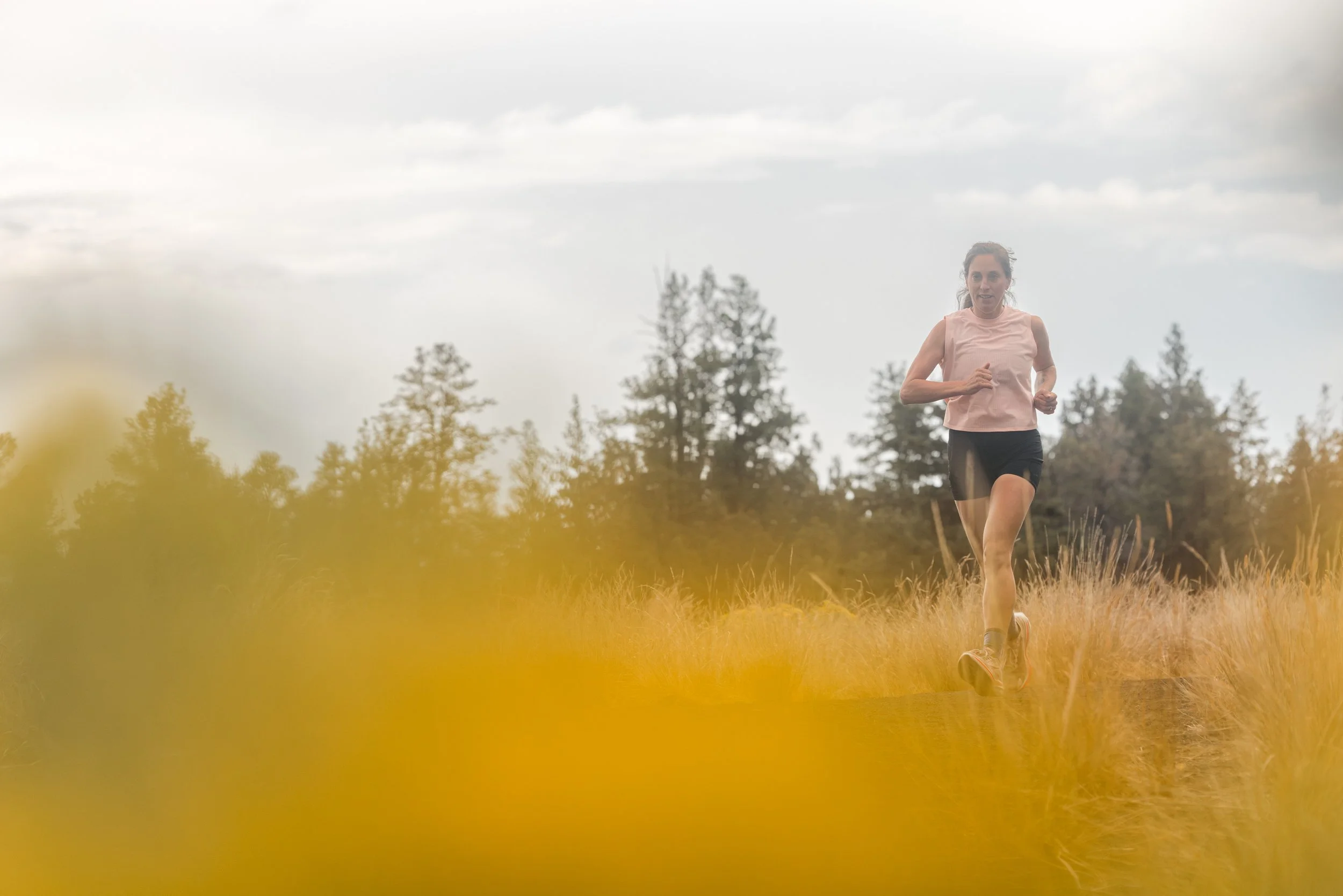 Woman running outdoors in a grassy field with trees in the background under a cloudy sky.