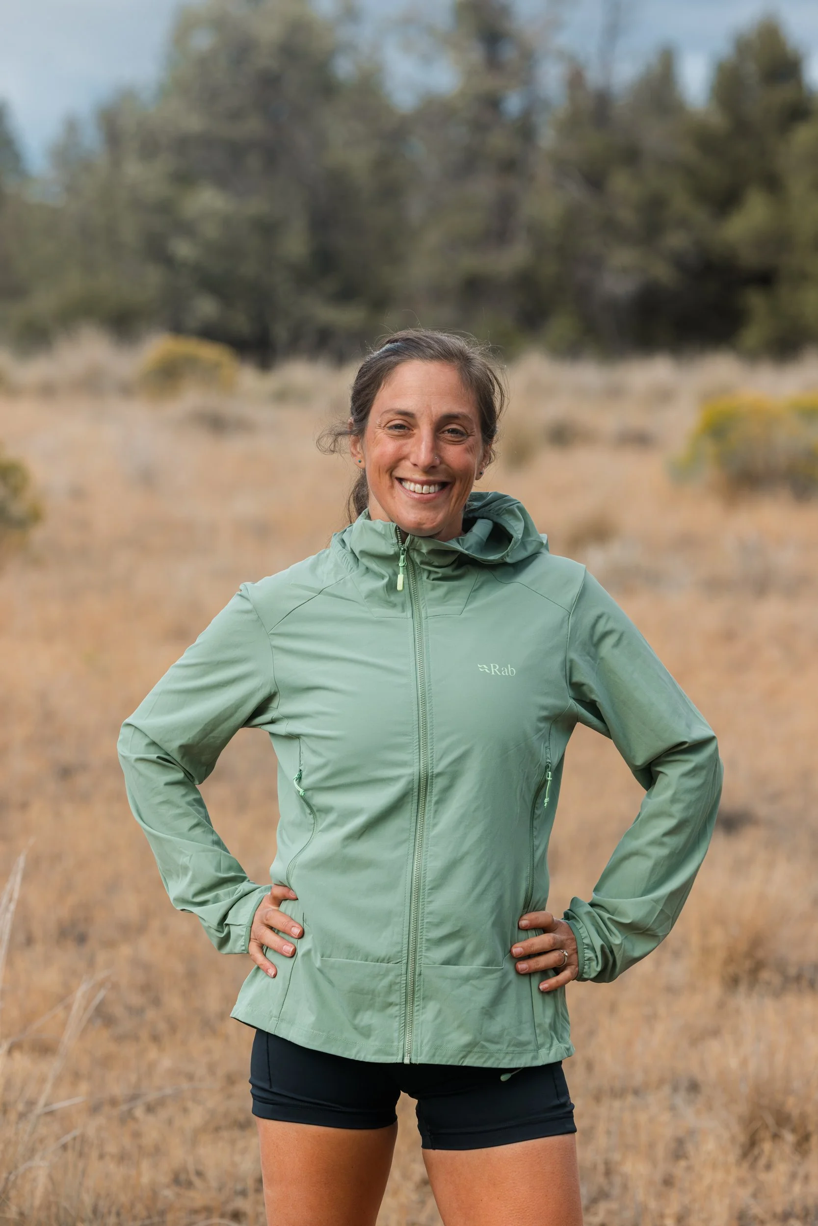 Smiling woman standing outdoors in a field with grassy and wooded background, wearing a light green jacket and black shorts.