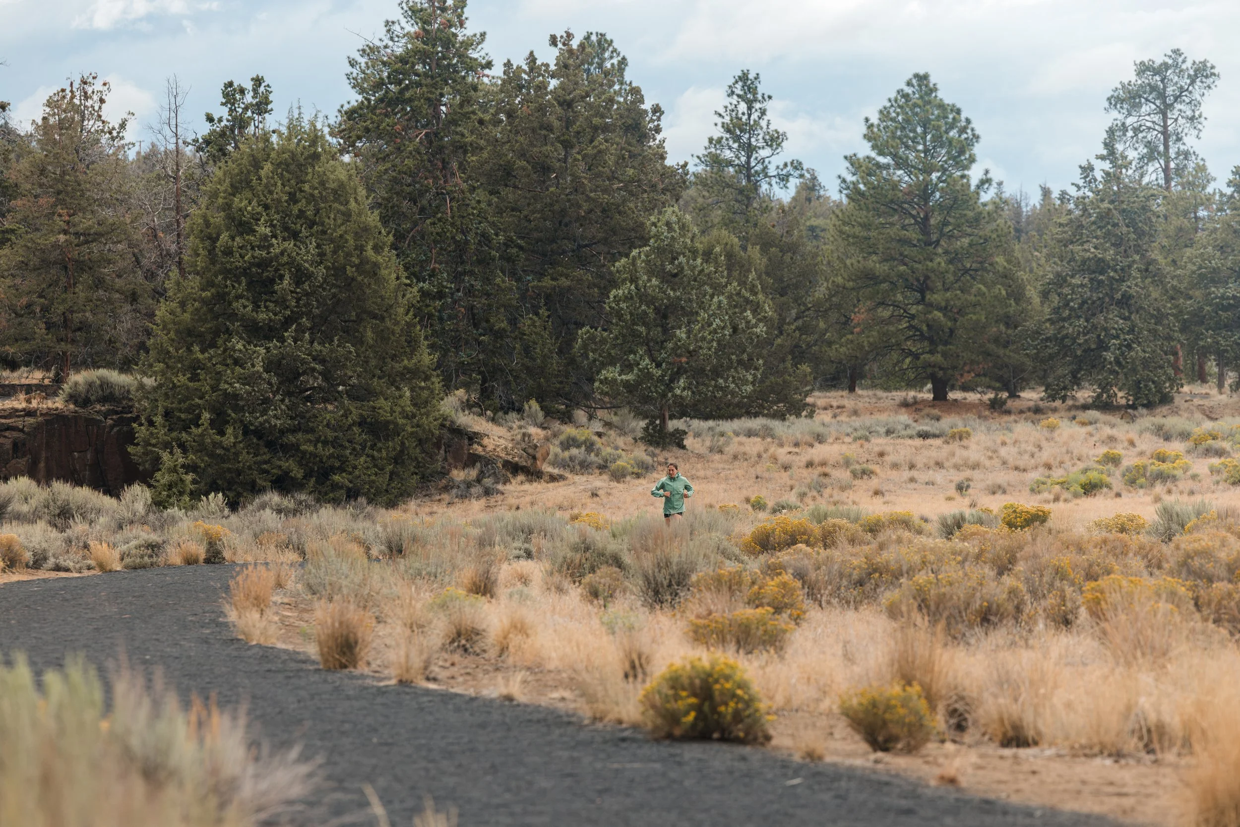 A person running in a field with dry grass and yellow wildflowers, surrounded by tall trees under a partly cloudy sky.