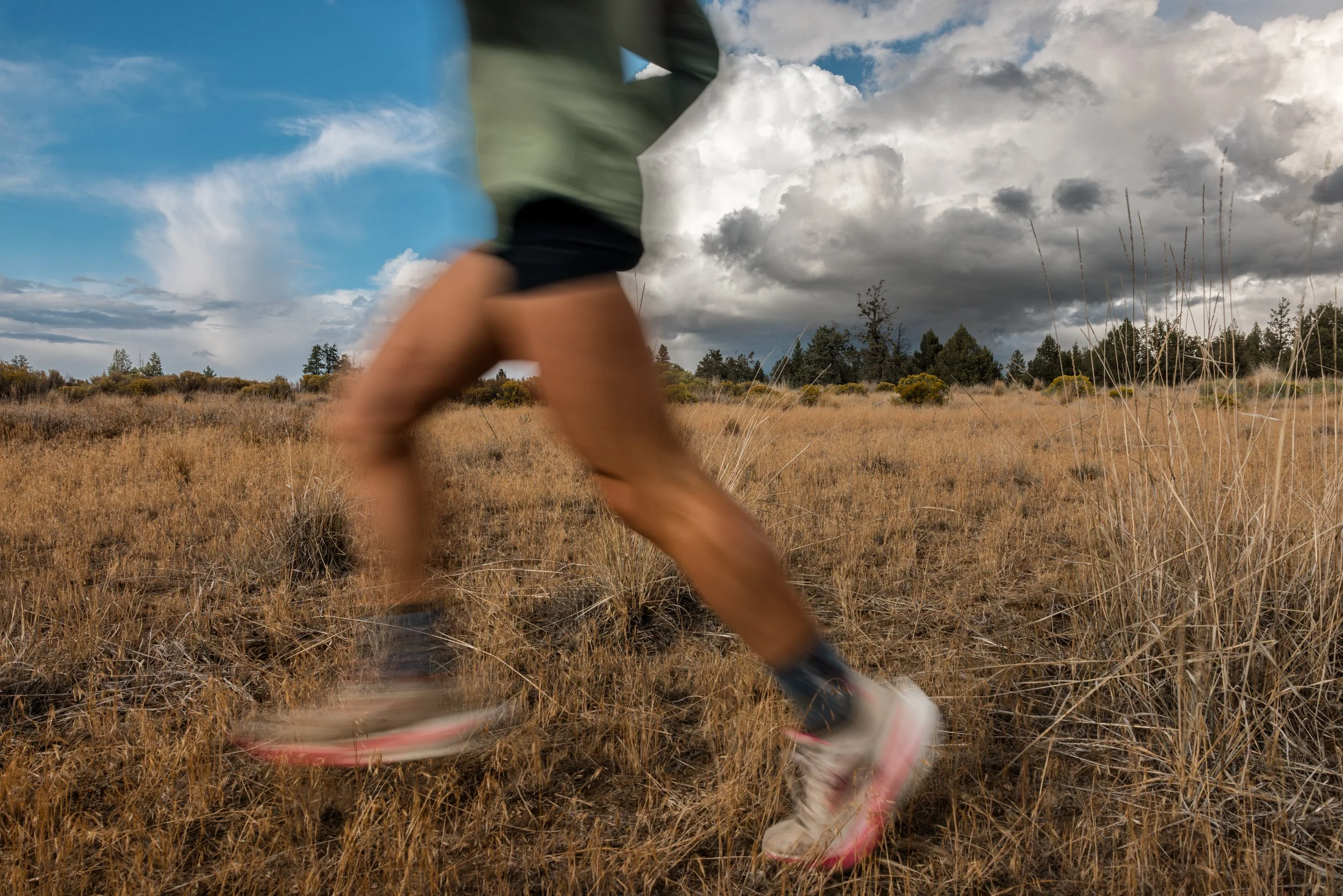 Blurred person rollerblading through a dry, grassy field on a partly cloudy day with dark clouds and blue sky.