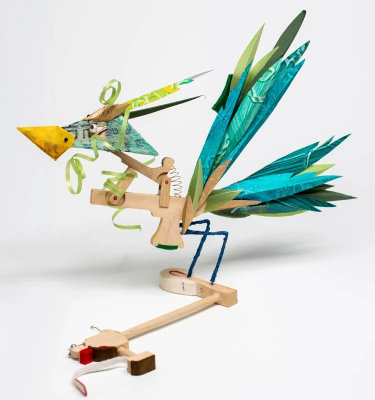 A colorful wooden and paper model of a mythical bird with a large beak, blue wings made of paper, and a long tail, set against a plain white background.