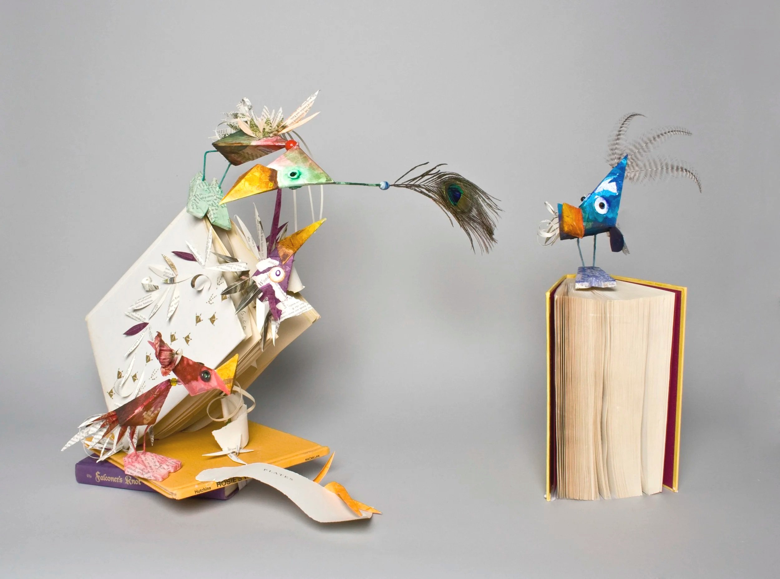 Colourful altered book sculpture made from recycled paper, depicting two birds with long beaks, feathers, and painted faces, positioned on book stacks against a plain gray background.