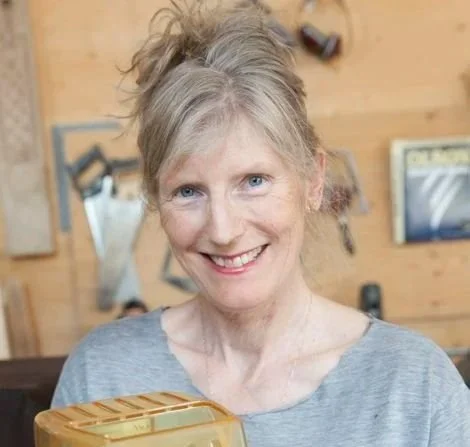 Smiling woman with short, light-colored hair and blue eyes, wearing a gray shirt, standing indoors with wooden walls in the background.