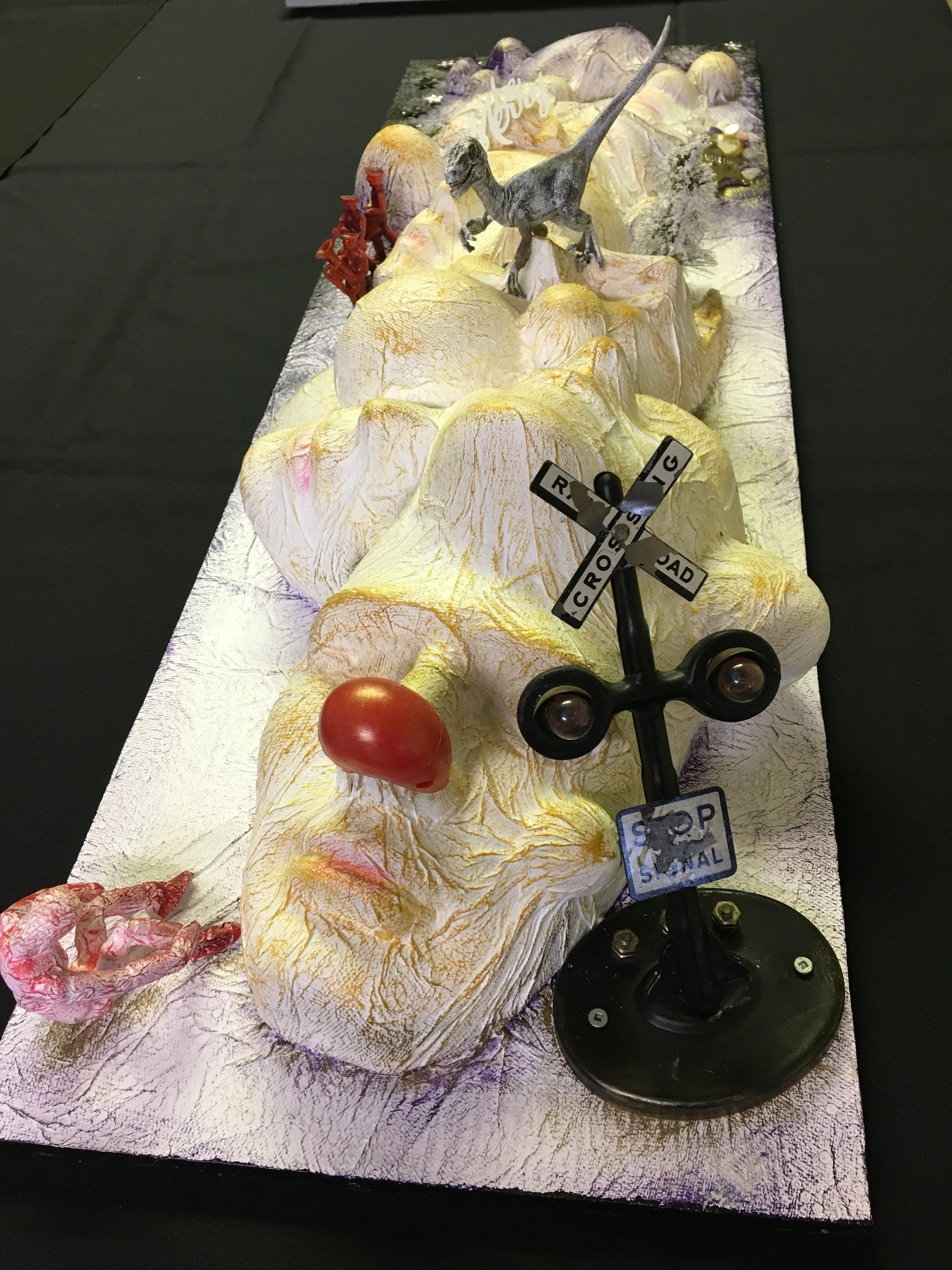 Sculpture designed with face masks to look like a landscape with yellow and purple mountains, a small dinosaur figurine, a red clown nose, and a miniature railroad crossing sign on a black surface.