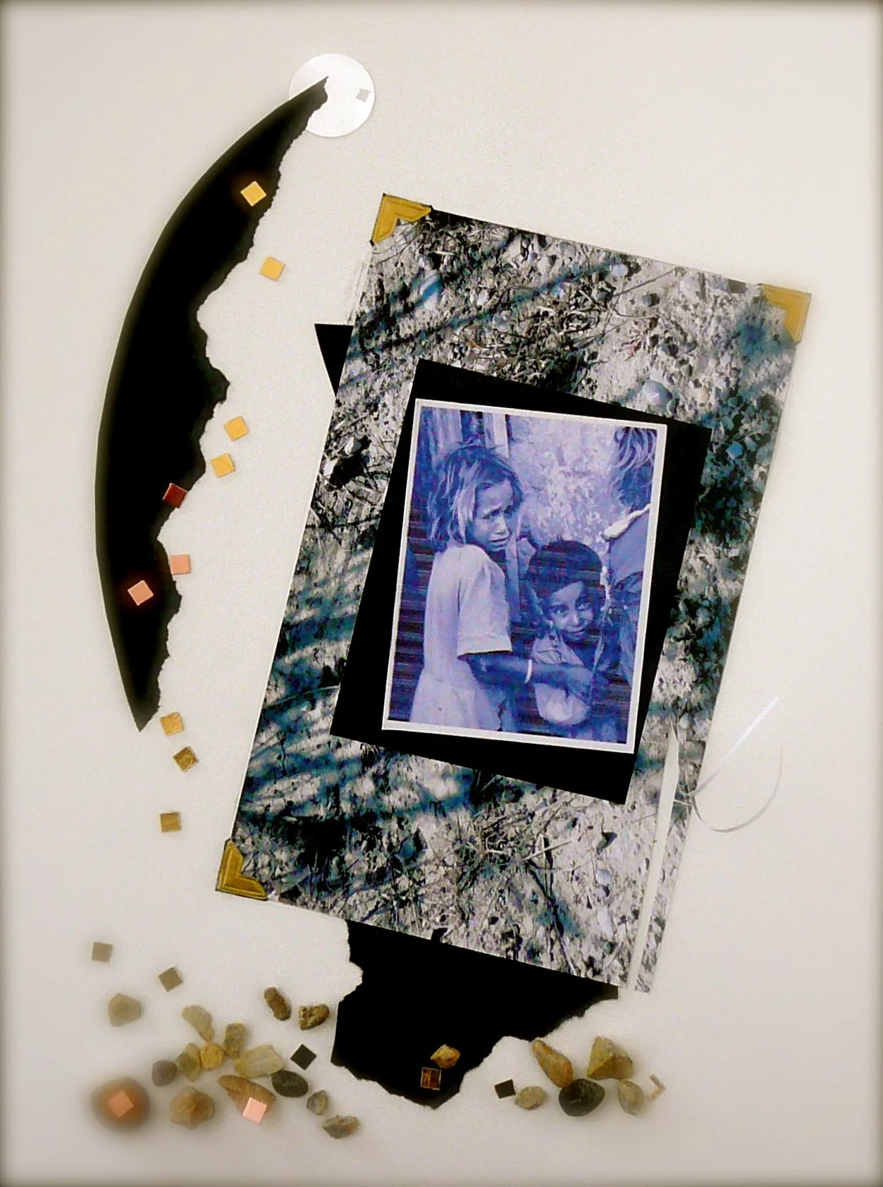 A mixed-media art piece with a black crescent shape, small square decorations, a black frame, a black and white textured border, and a central blue-tinted photograph of two children, with small rocks and colorful squares scattered around on a white s