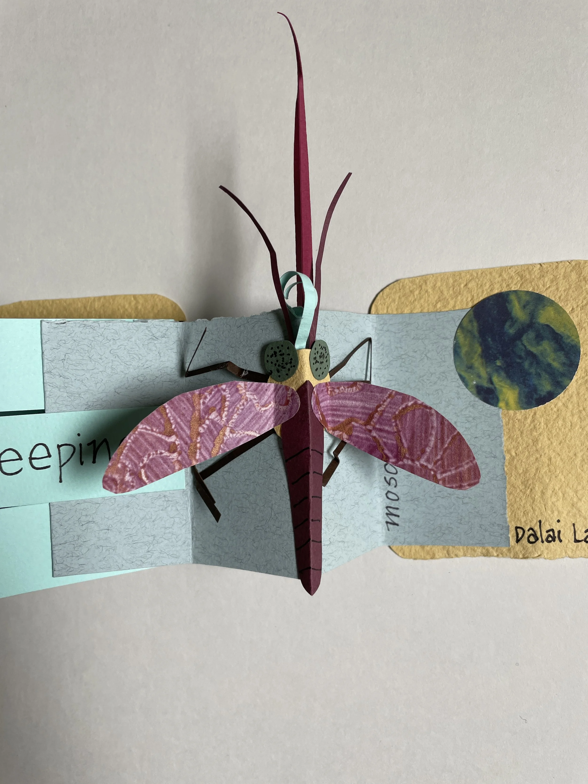 Paper craft of a butterfly with pink and brown wings, green spots on its body, and pink antennae, attached to a light blue background with handwritten words.
