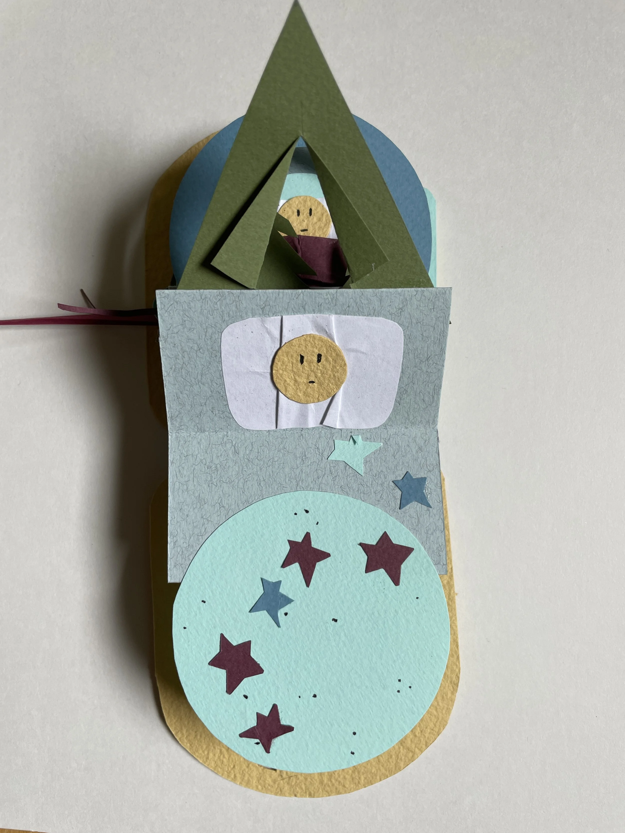 Colorful paper craft featuring a star pattern, a face drawing on a moon, and a spaceship or rocket shape with a character inside.