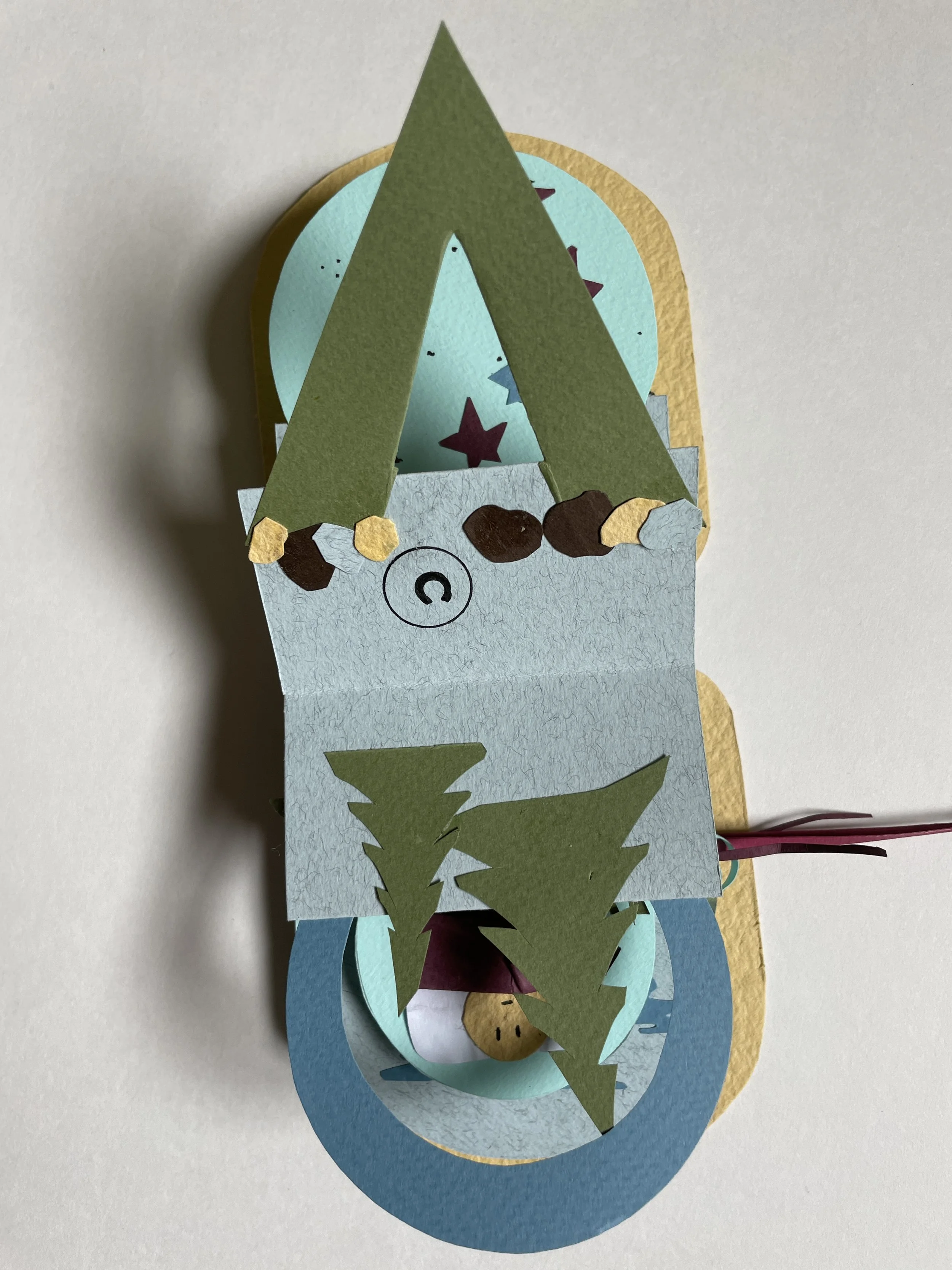 Colorful 3D paper craft of a Christmas scene featuring a pine tree, decorated with stars, and a small bear figure at the bottom, with layers of paper in blue, green, and yellow.