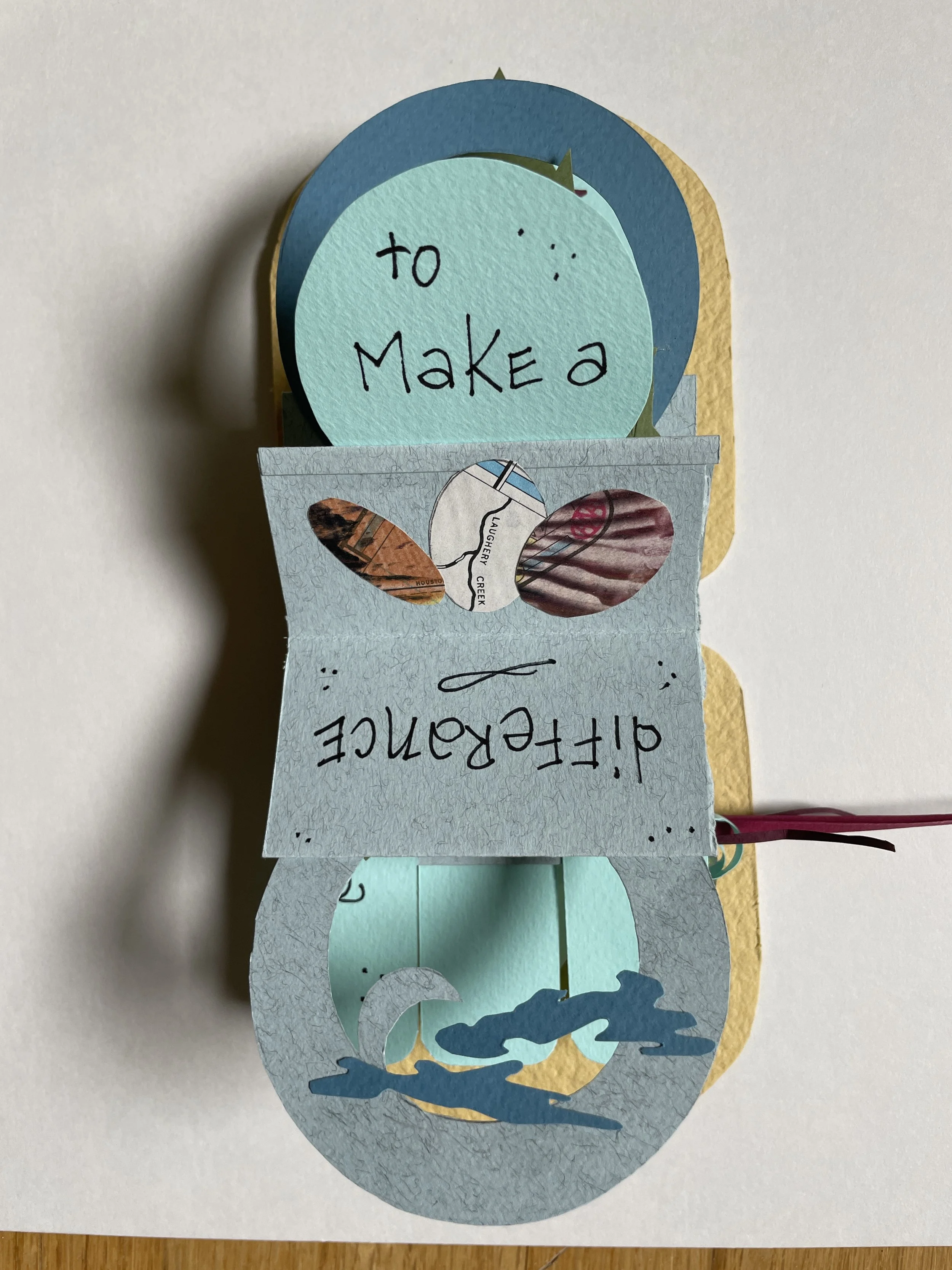 A handmade paper craft with a blue ocean wave and a dolphin, featuring a circular note that says 'to MAKE a' and cutouts with words 'Laughter Creek' and some illustration of ocean waves.