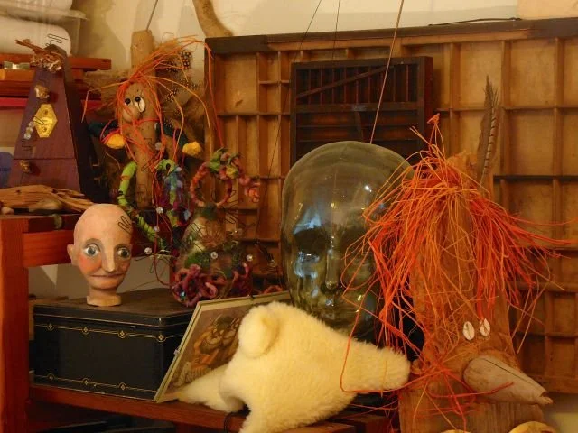 Display of Halloween or horror-themed decorations including a large glass skull, a white plush mouse, creepy doll head, colorful puppets, and orange fiber decorations on a wooden surface.