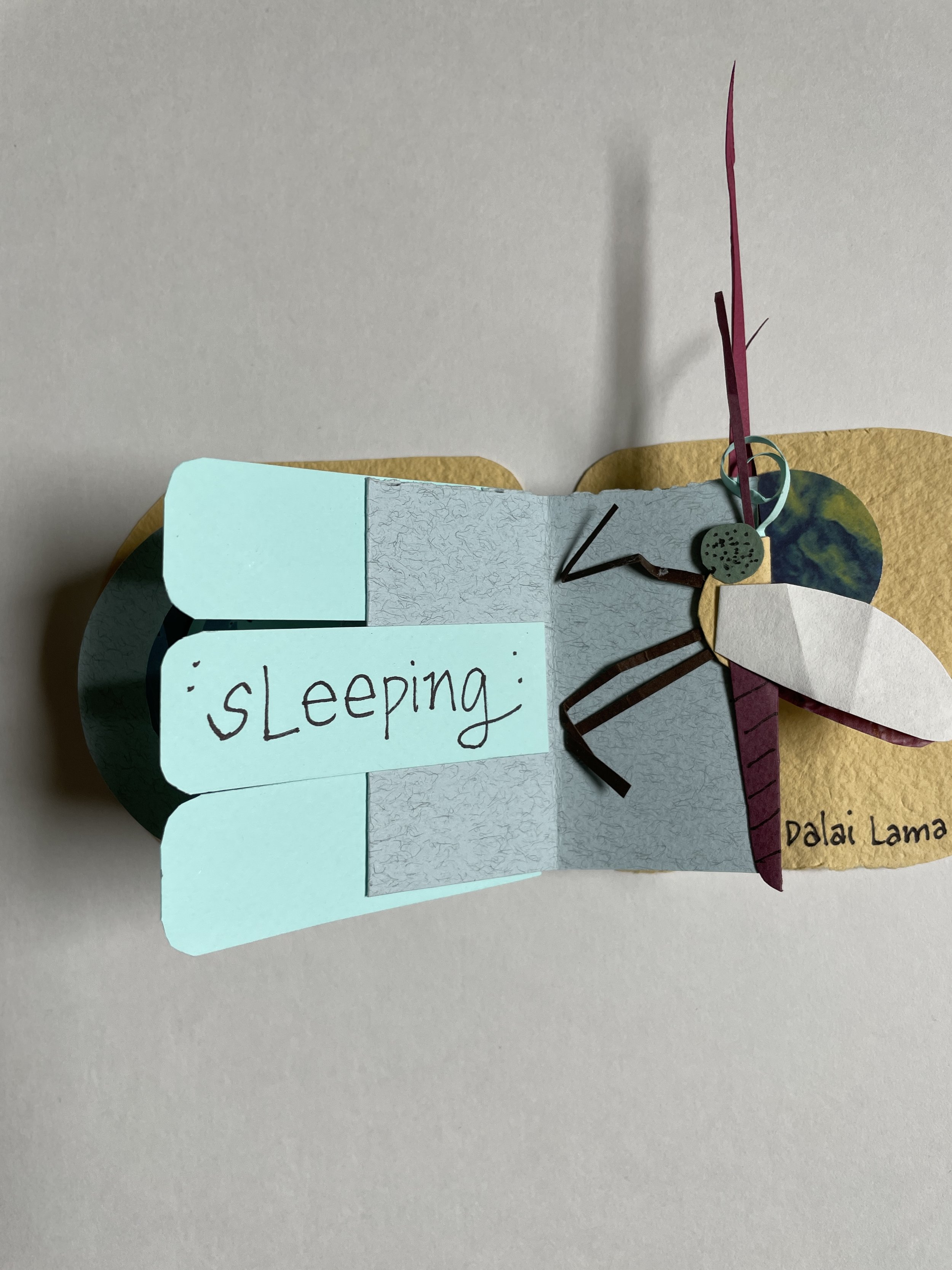 A handmade paper craft of a dragonfly. The dragonfly is made with cut-out paper in various colors and textures, attached to a yellow background. There is a small handwritten note in blue paper that says 'sleeping' connected to the craft. The craft ap