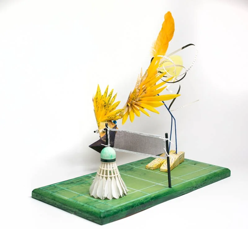 Whimsical sculpture of a badminton player with butterfly wings on a green base.