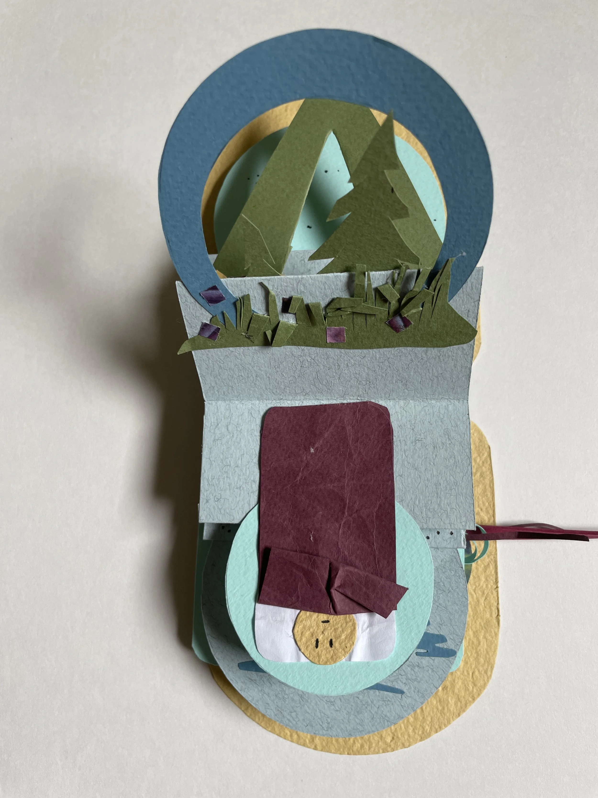 A layered paper craft of an animal, possibly a dog or a bear, lying on a circular bed with a yellow, green, and blue background, decorated with grass and small flowers.