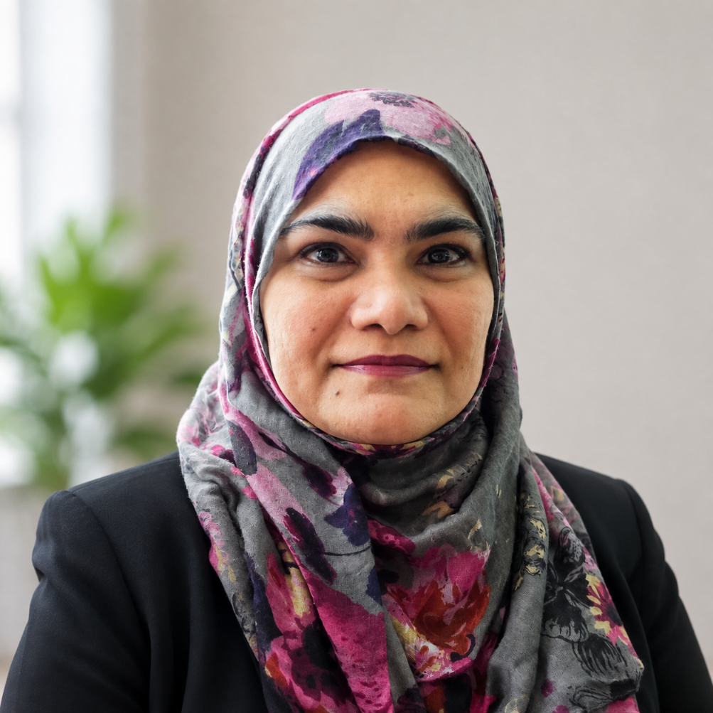 A woman wearing a colorful floral hijab and a black blazer, smiling at the camera with a blurred green plant in the background.