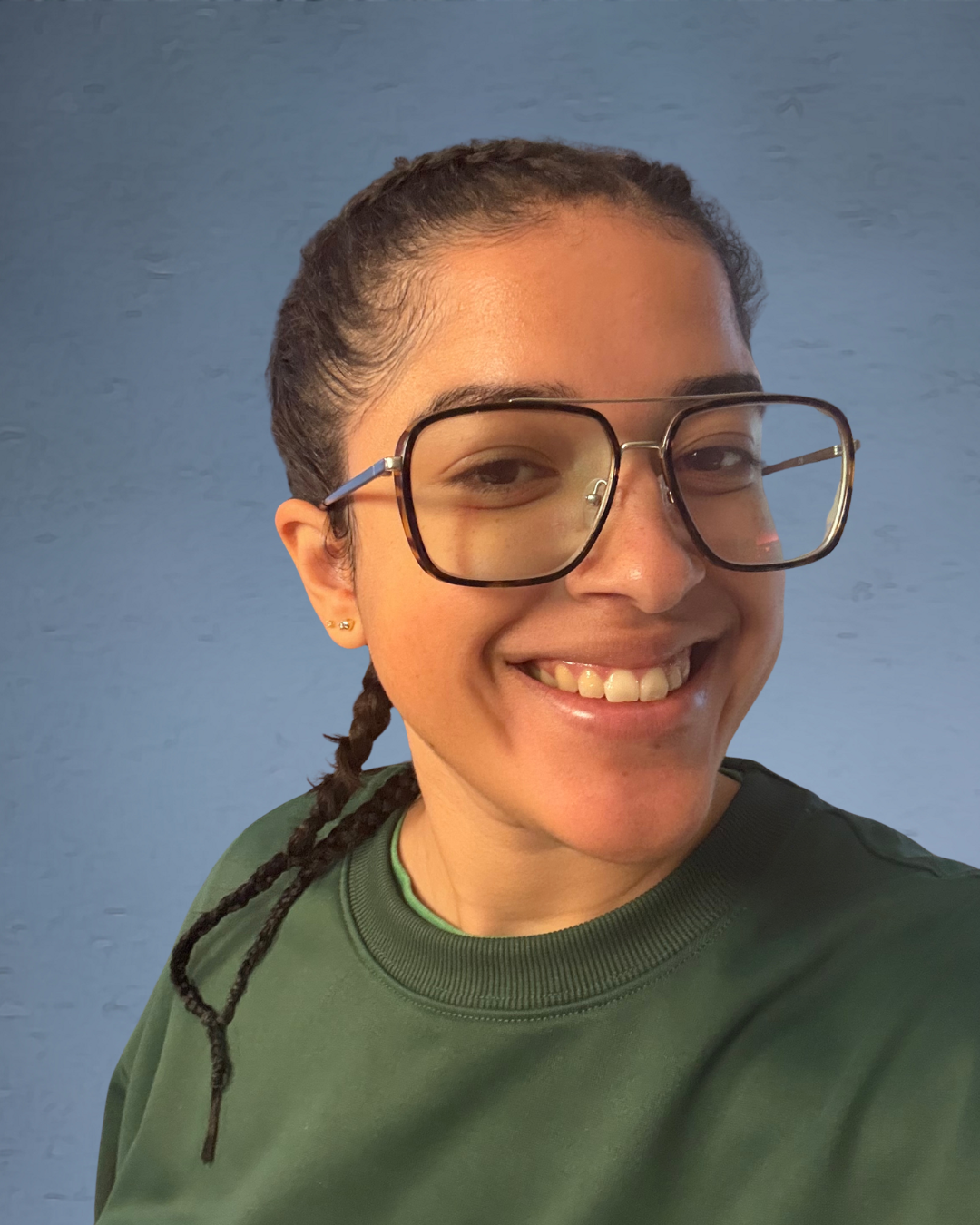 A smiling woman with glasses, dark hair tied in a braid, wearing a green shirt against a blue wall.