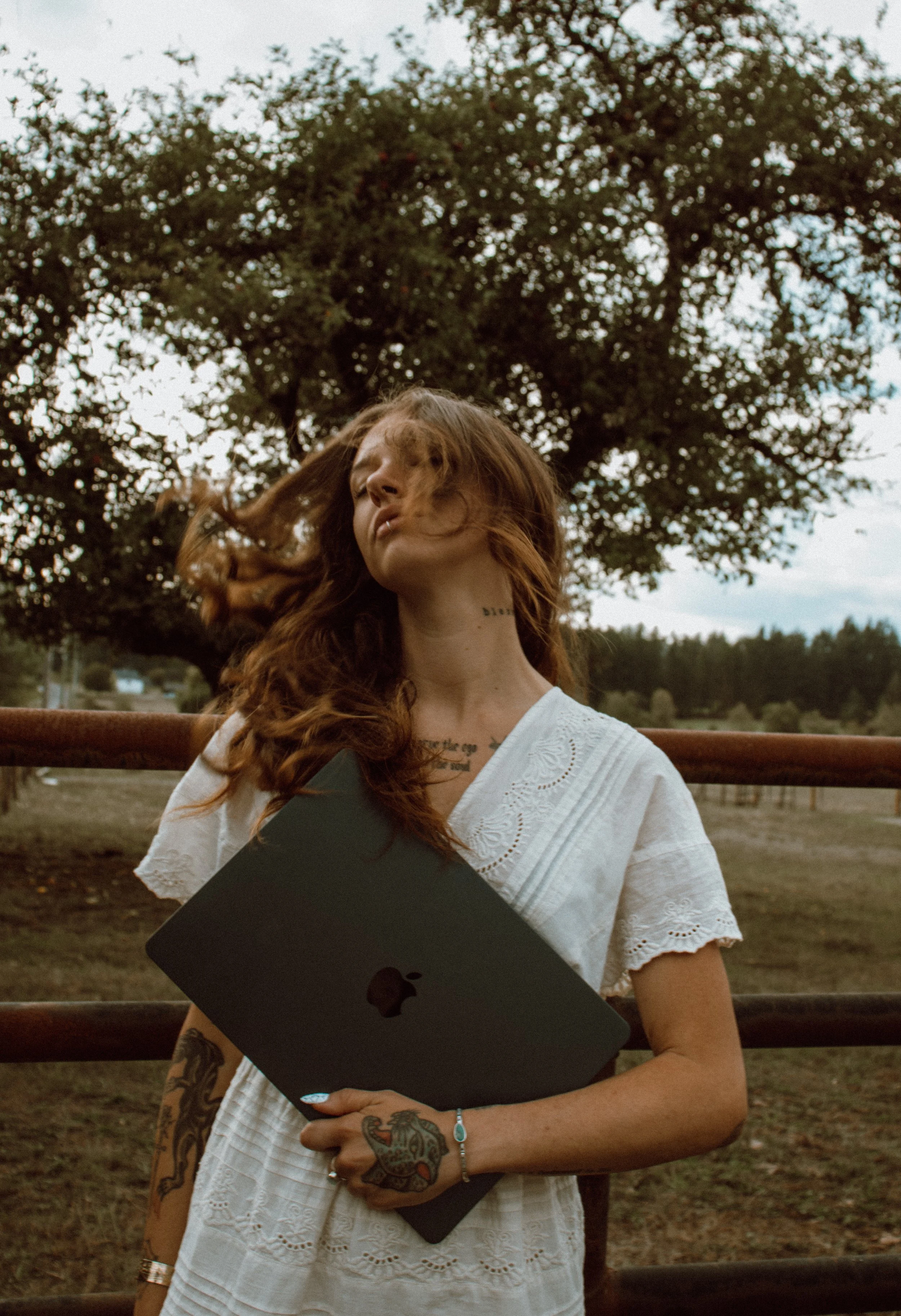 A woman with long red hair and tattoos on her arm and neck, wearing a white embroidered dress, holding a closed laptop, standing outdoors in front of a wooden fence, with a large tree and distant landscape under a cloudy sky.