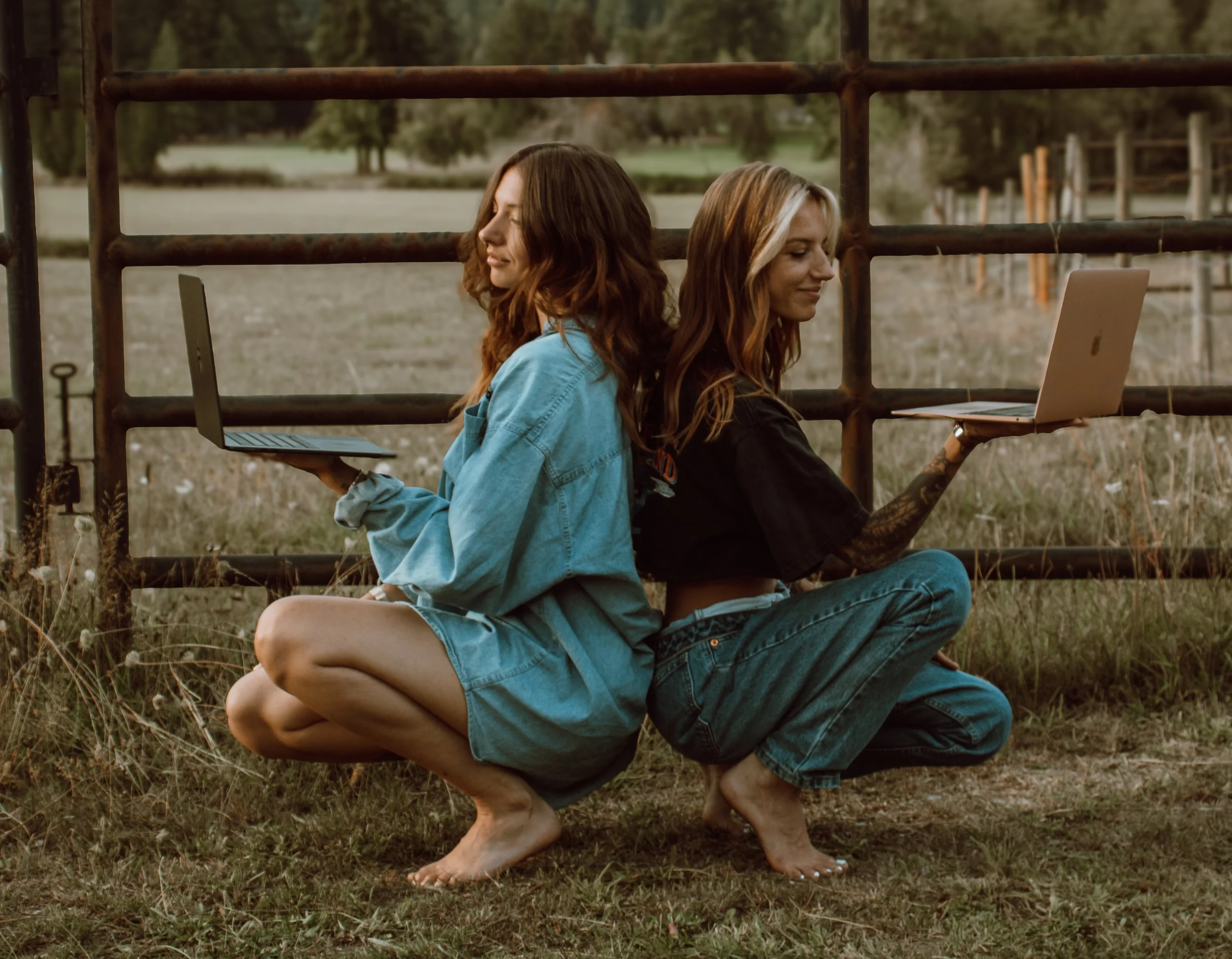 Two women sitting back-to-back outdoors on grass, each holding a laptop, with a rural background and a metal fence.