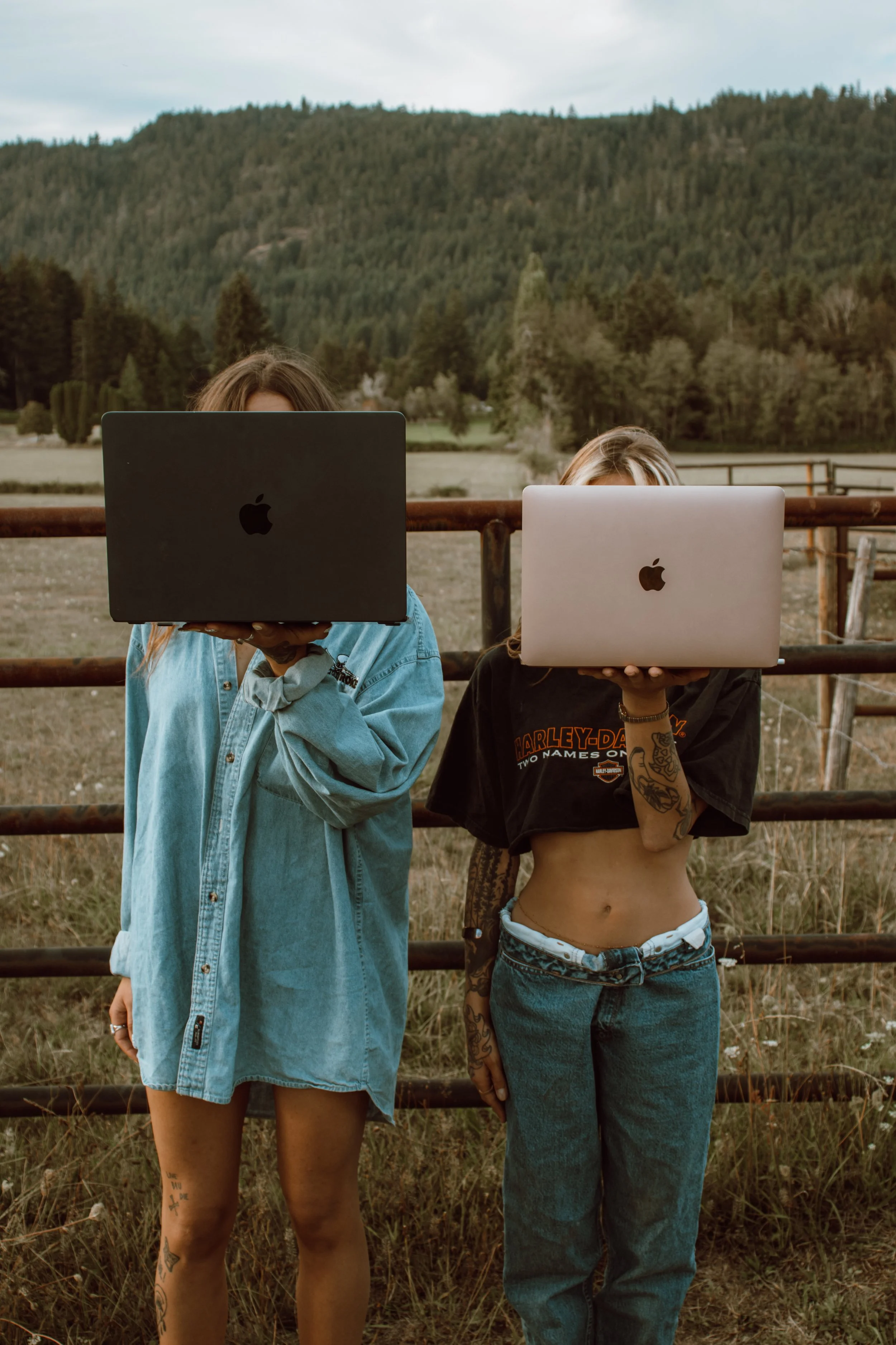 Two women standing outdoors behind a wooden fence, holding up laptops to cover their faces. One wears an oversized blue denim shirt, the other a black Harley-Davidson t-shirt and jeans. The scene is set in a rural area with grassy fields and a forested hill in the background.