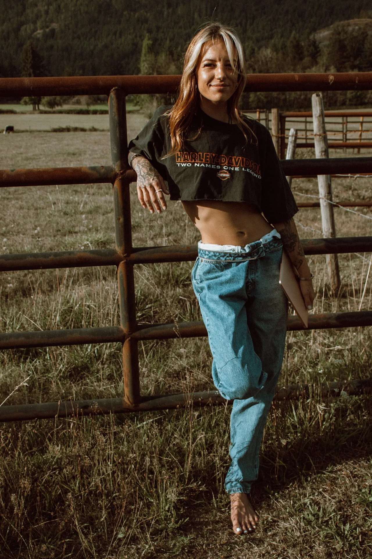A young woman with long reddish hair, tattoos on her hand, wearing a black Harley-Davidson T-shirt, loose blue jeans, and barefoot, standing outdoors by a rusty metal fence in a rural field with trees and hills in the background, during sunset.