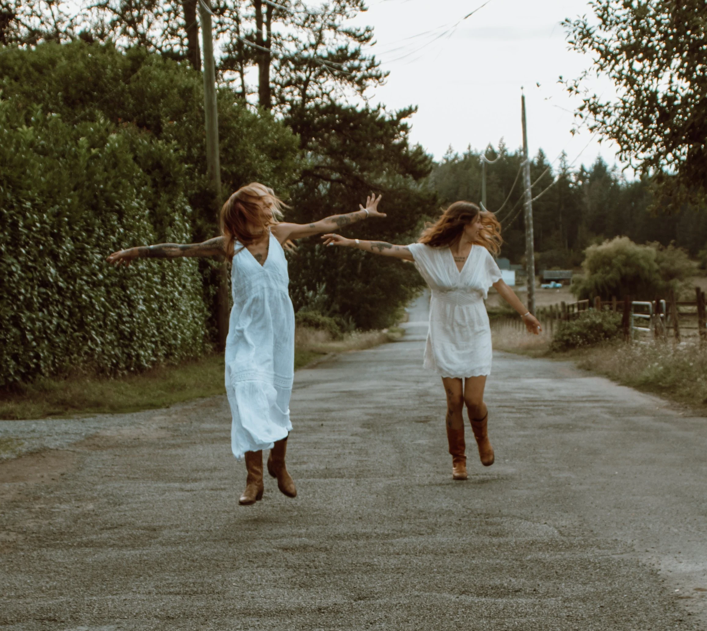 Two women in white dresses and brown boots dancing on a dirt road surrounded by green bushes and trees during daytime.