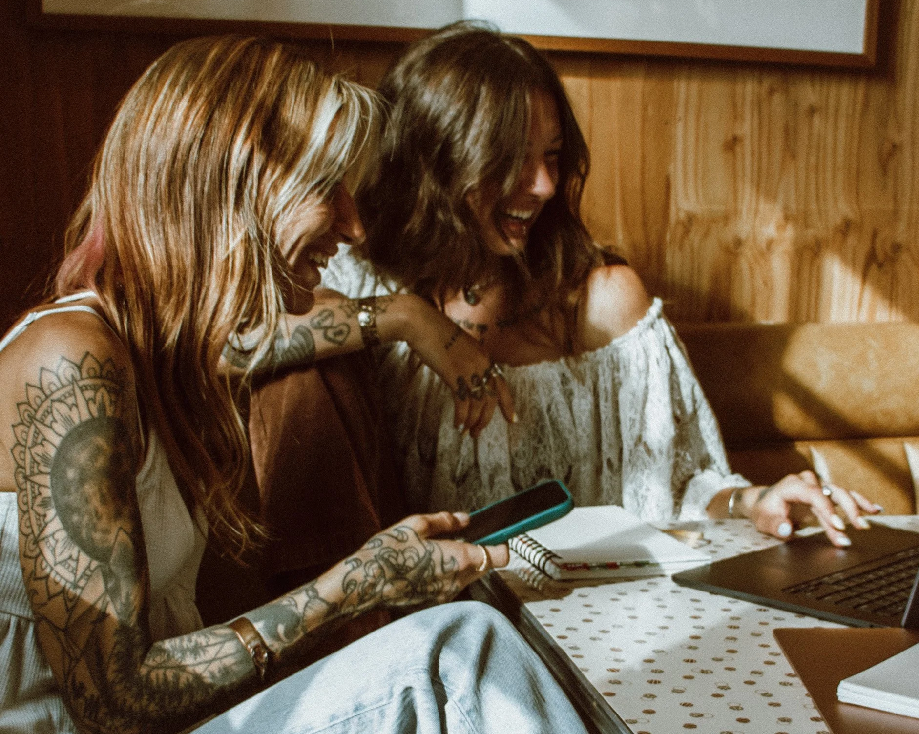 Two women with tattoos sitting at a table, laughing and looking at a phone together in a cozy wooden-paneled room.