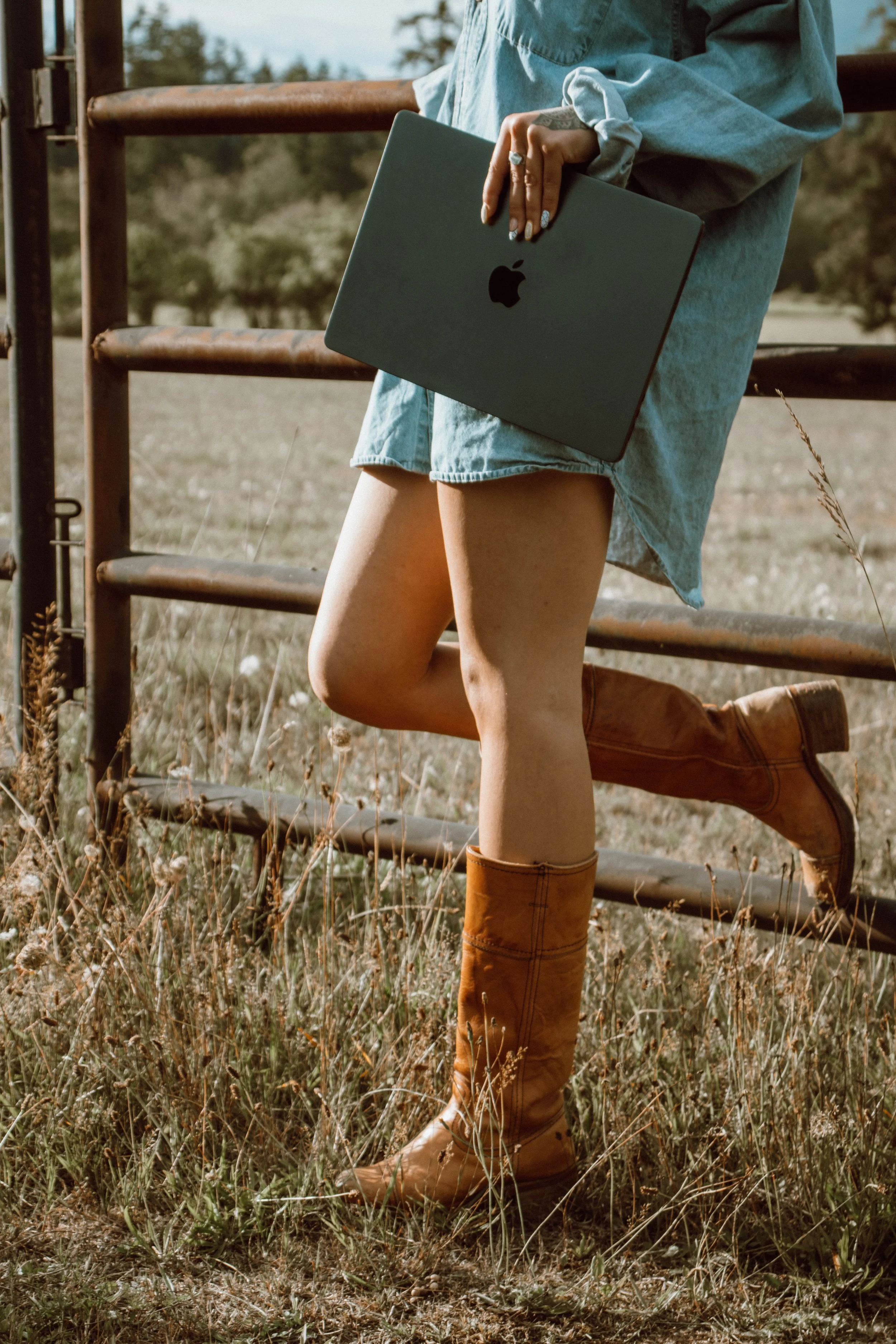 Person holding a closed gray MacBook with a black apple logo, wearing a denim dress and brown boots, standing outdoors on grass with a rusty metal fence in the background.