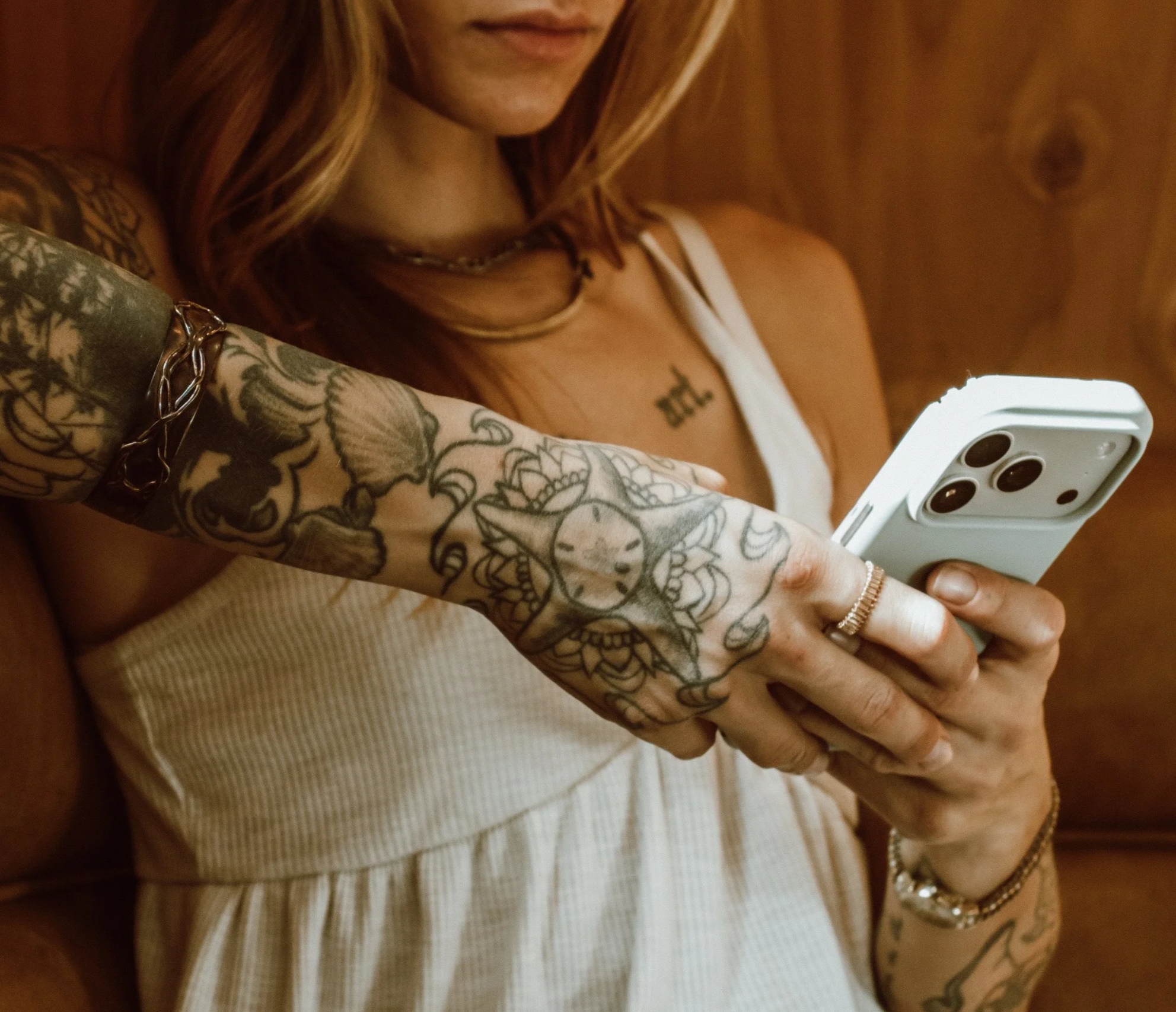 A woman with tattoos on her arm, wearing a white sleeveless top, holding a smartphone with a triple camera, looking at the screen. She has necklaces and a bracelet, with a wooden wall in the background.
