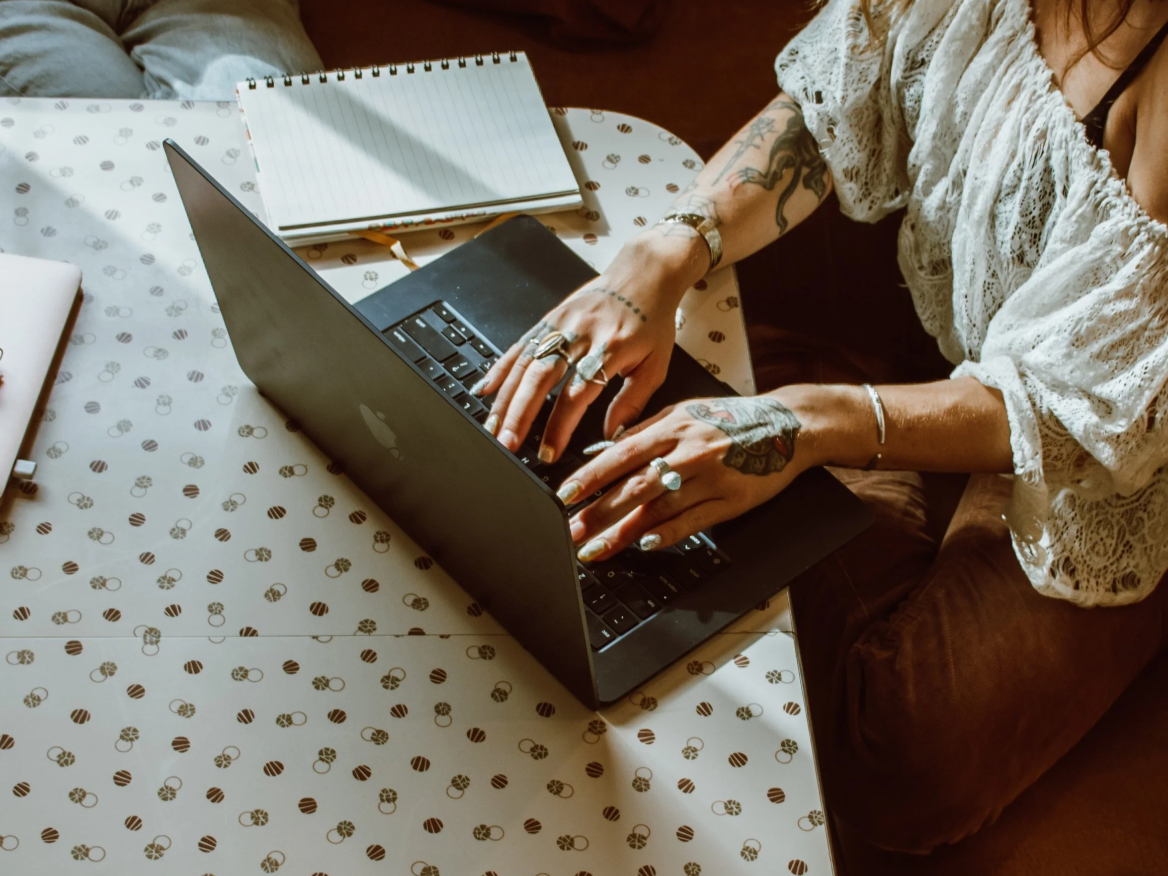 A person with tattoos on their arms, wearing rings and a bracelet, typing on a laptop. They are seated at a table with a notepad and a closed laptop, wearing a white lace blouse and brown pants.
