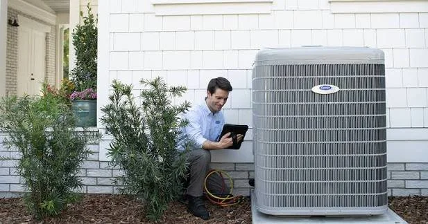 A man crouching next to an air conditioning unit outside a house, holding a tablet device.