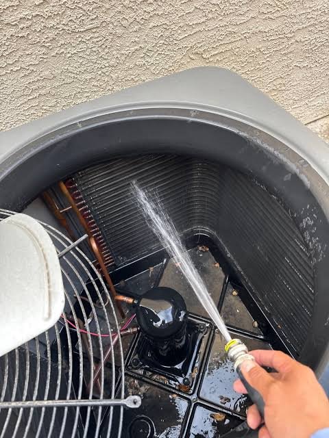 A person is cleaning an outdoor air conditioning unit with a garden hose, spraying water inside the unit.